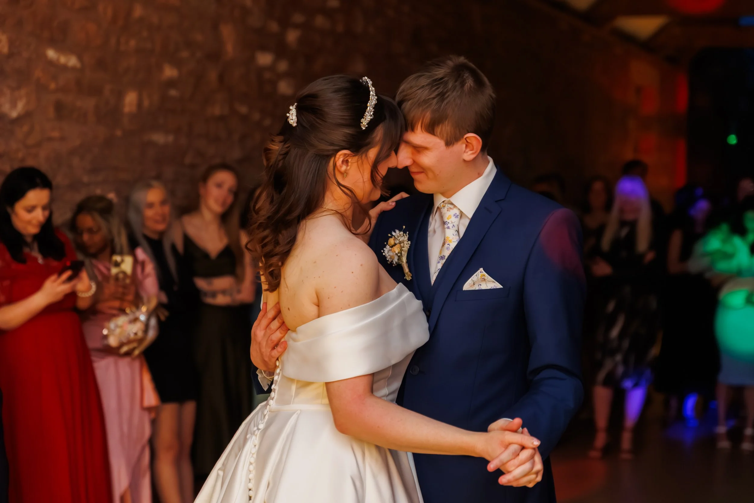 A bride and groom dance closely at their wedding reception, surrounded by guests. The bride has dark hair with decorative hairpieces, and the groom wears a blue suit with a floral tie.