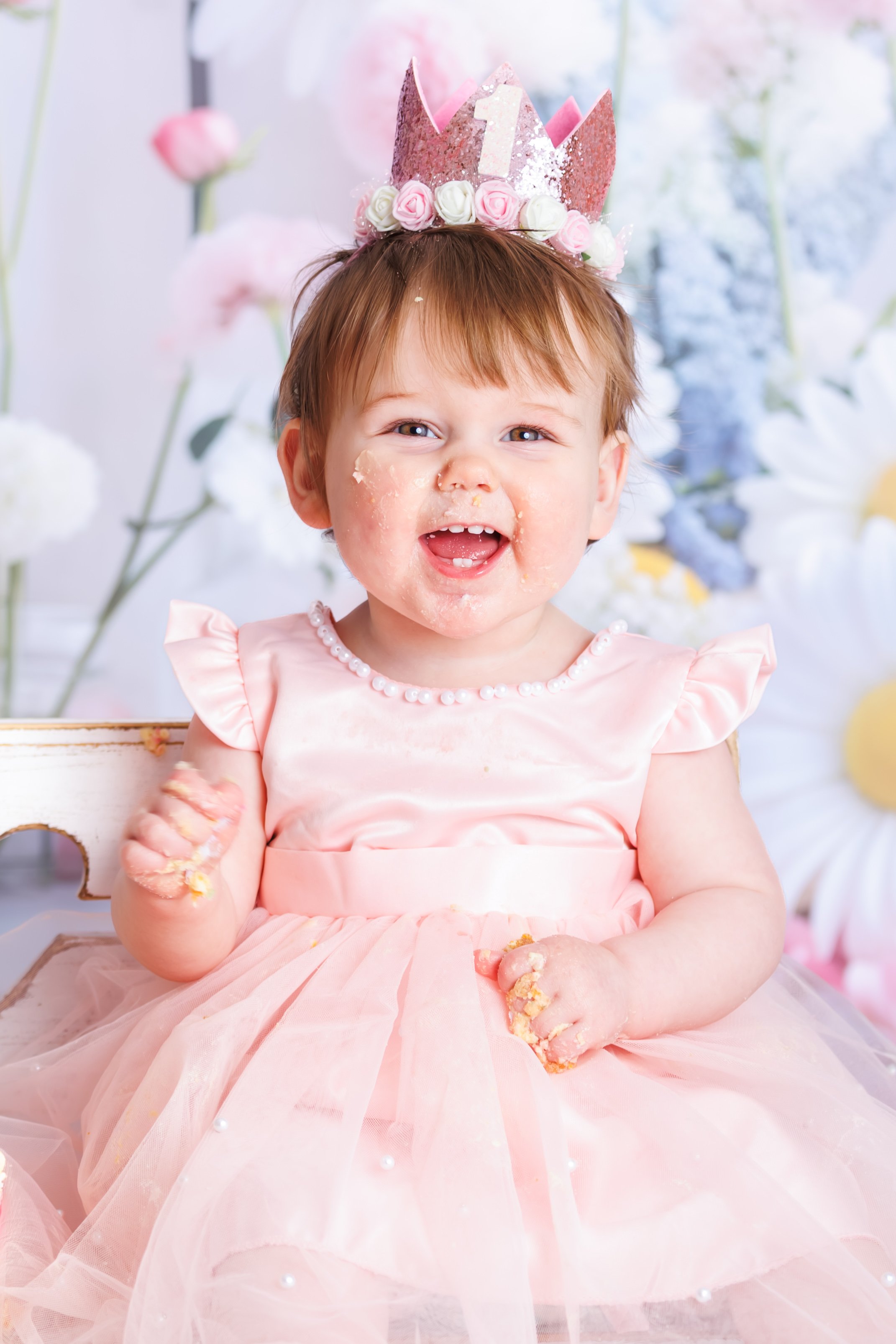 A young girl with red hair wearing a pink dress and a pink birthday crown with the number 1 on it. She has cake on her face and hands, and is smiling.