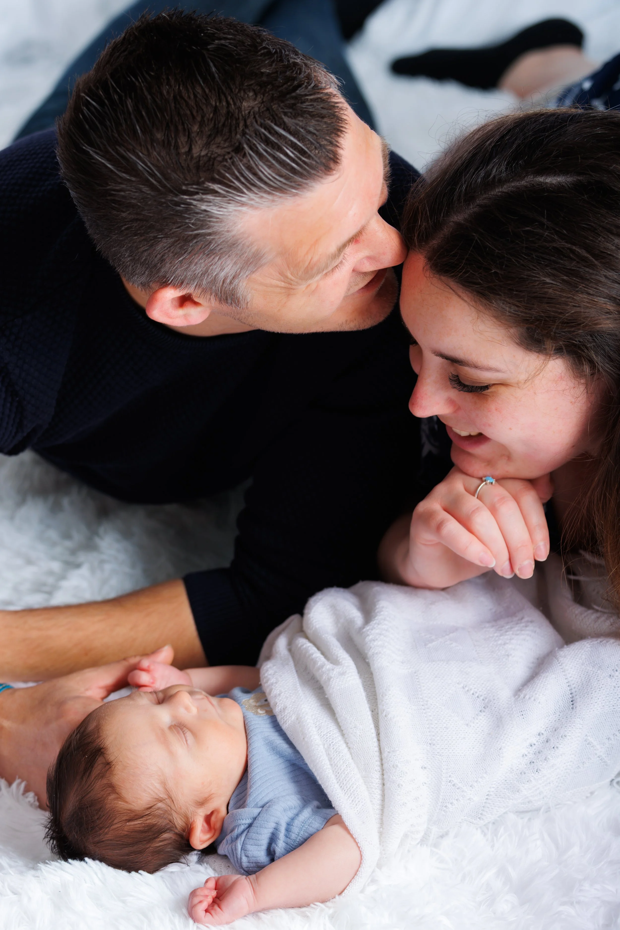 A man leaning over a baby, smiling and holding the baby's hand, while the baby lies on a fluffy white blanket and looks up at the man.