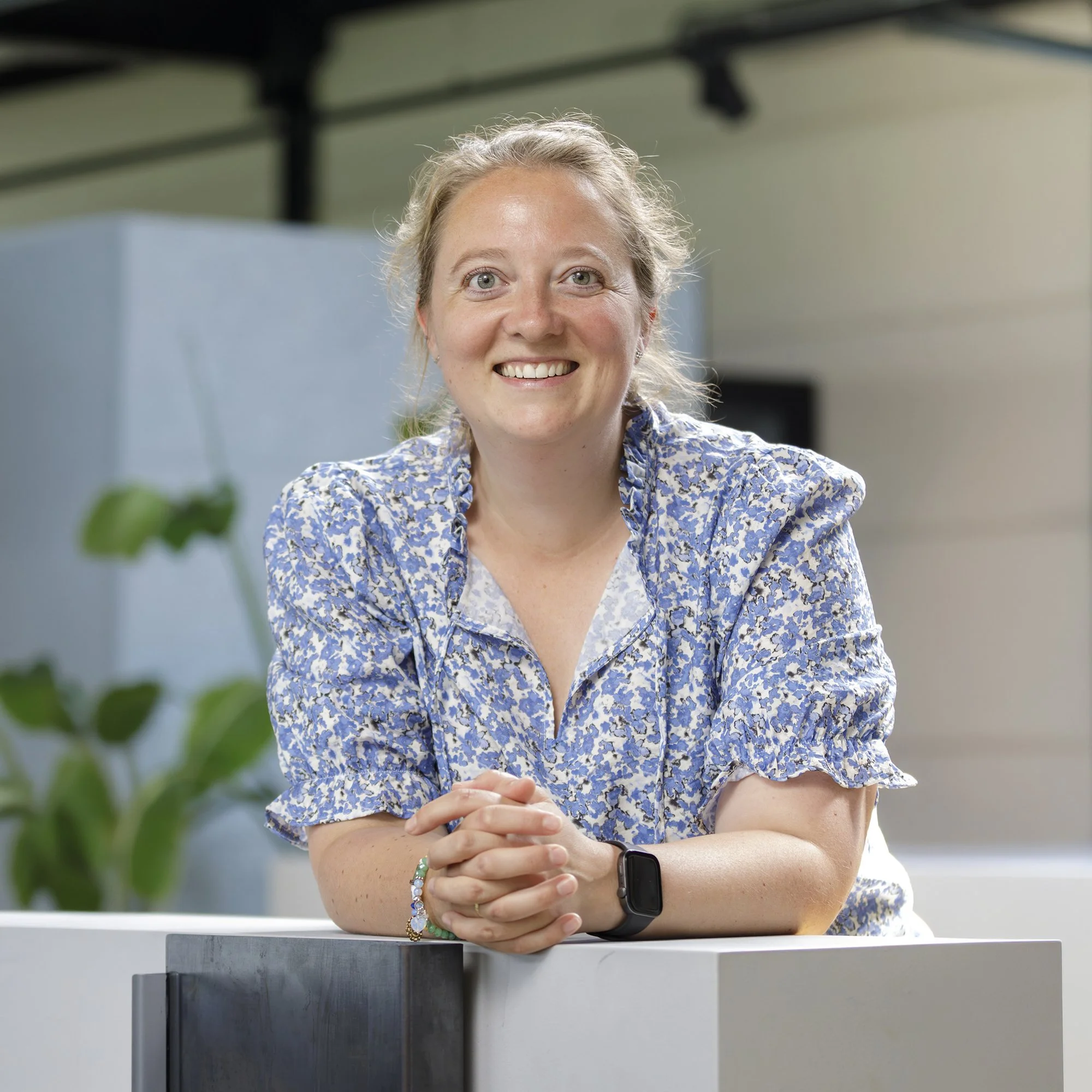 Een vrolijke vrouw met een grijs haar, glimlachend en zittend met haar handen gevouwen op een tafel, in een moderne kantooromgeving met groene planten en zwarte metalen plafondbalken.