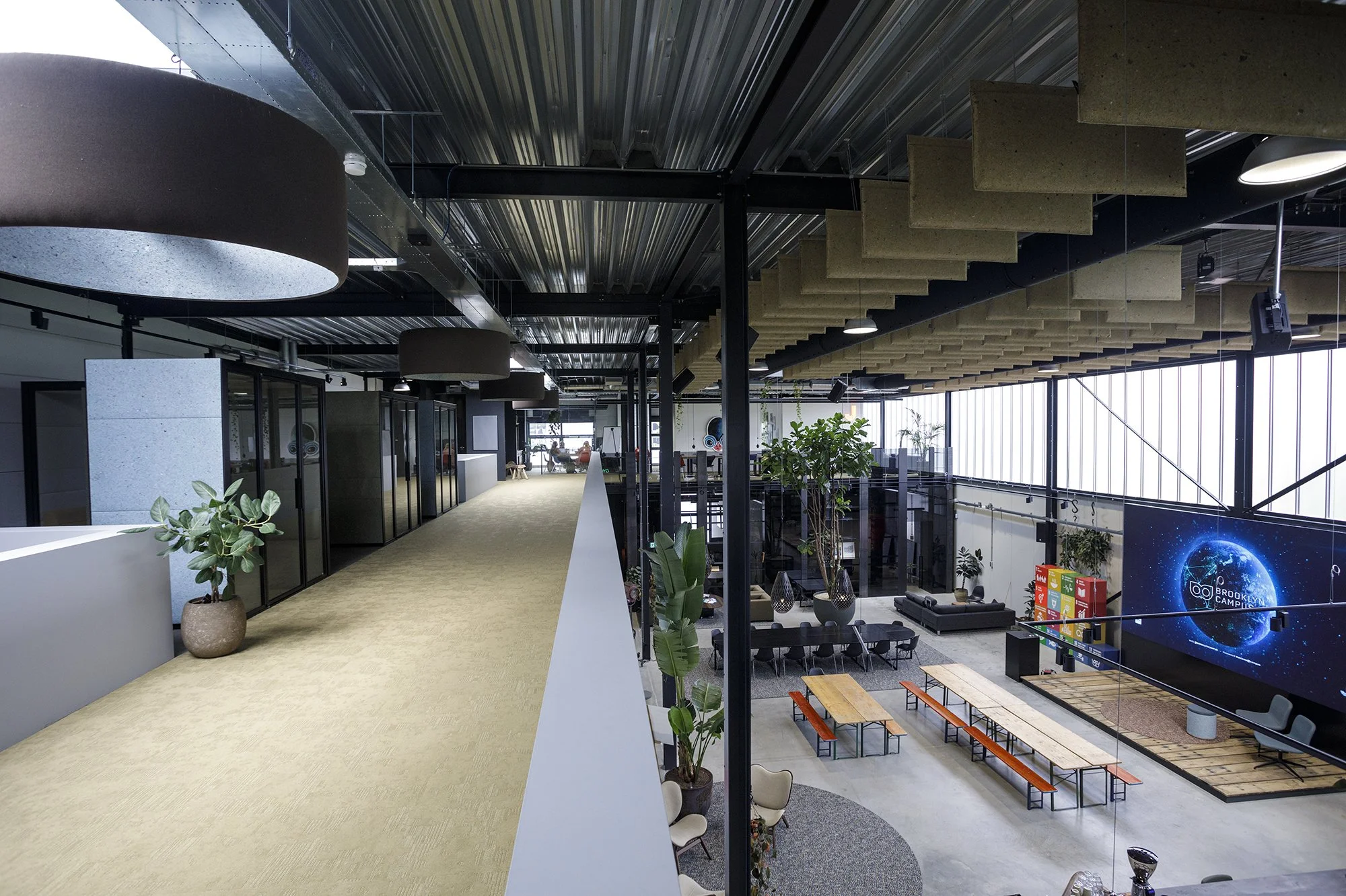 Interieur van een modern kantoorpand met een open werkruimte, planten, zitgedeeltes en een groot scherm met een wereldkaart en de tekst 'Brooklyn Camp'.