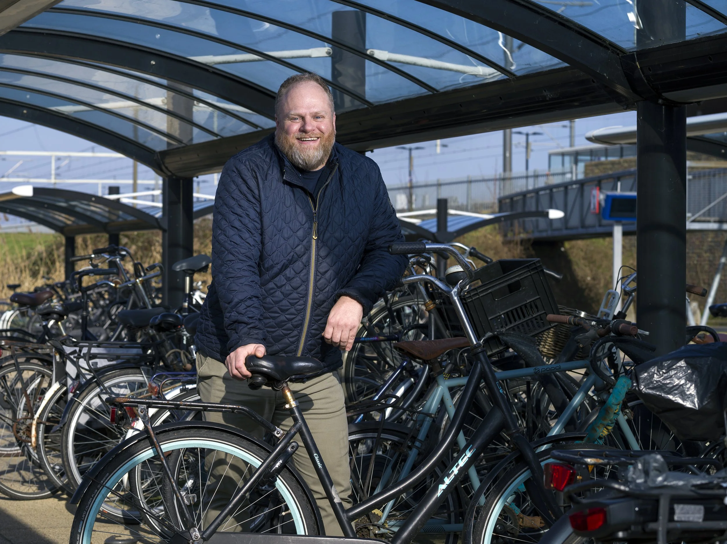 Lachende man met een baard die op een fiets rust onder een glazen stallingskap, omringd door meerdere fietsen, op een fietsparkeergelegenheid buiten.