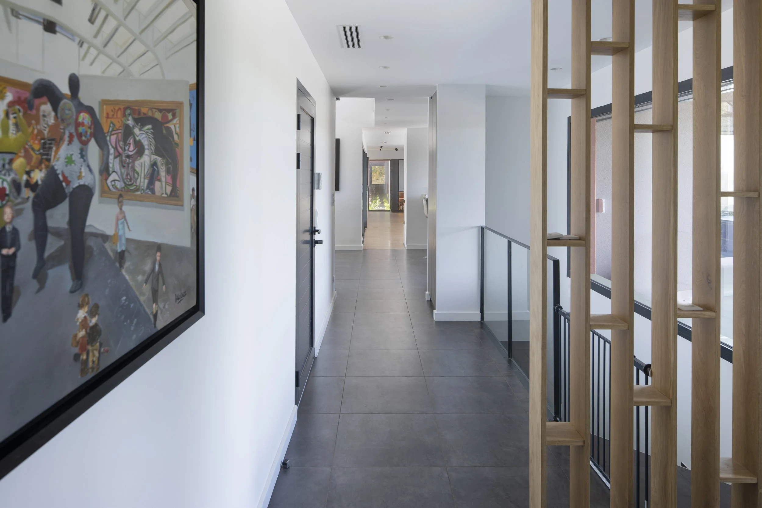 Couloir dans un bâtiment moderne avec peinture et décoration minimaliste, murs blancs, sol carrelé gris foncé, éléments en bois naturel, porte en métal, fenêtres laissant entrer de la lumière naturelle.