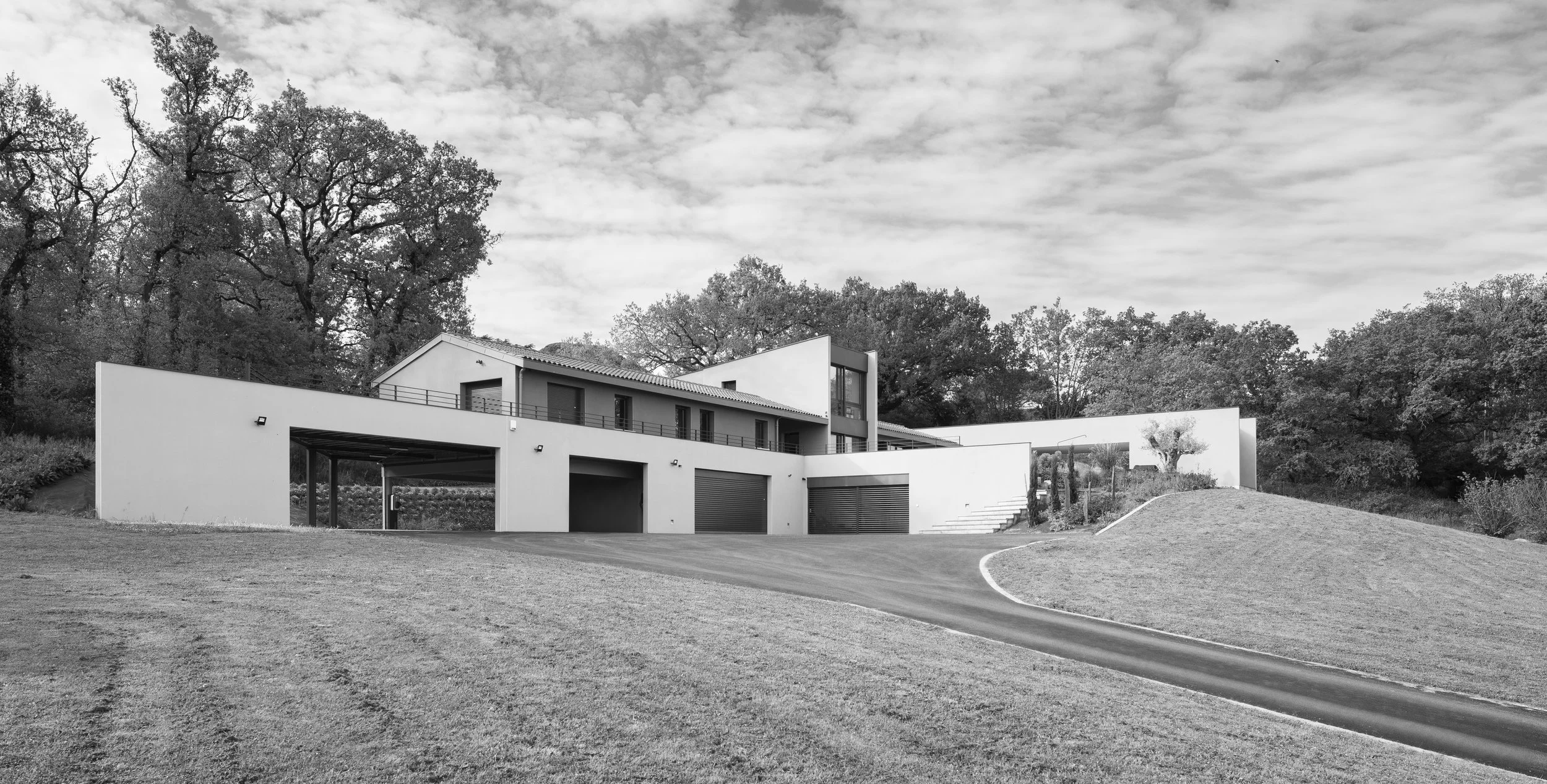 Maison moderne avec plusieurs niveaux, garages, et un chemin serpentant à travers une pelouse, entourée d'arbres, en noir et blanc.