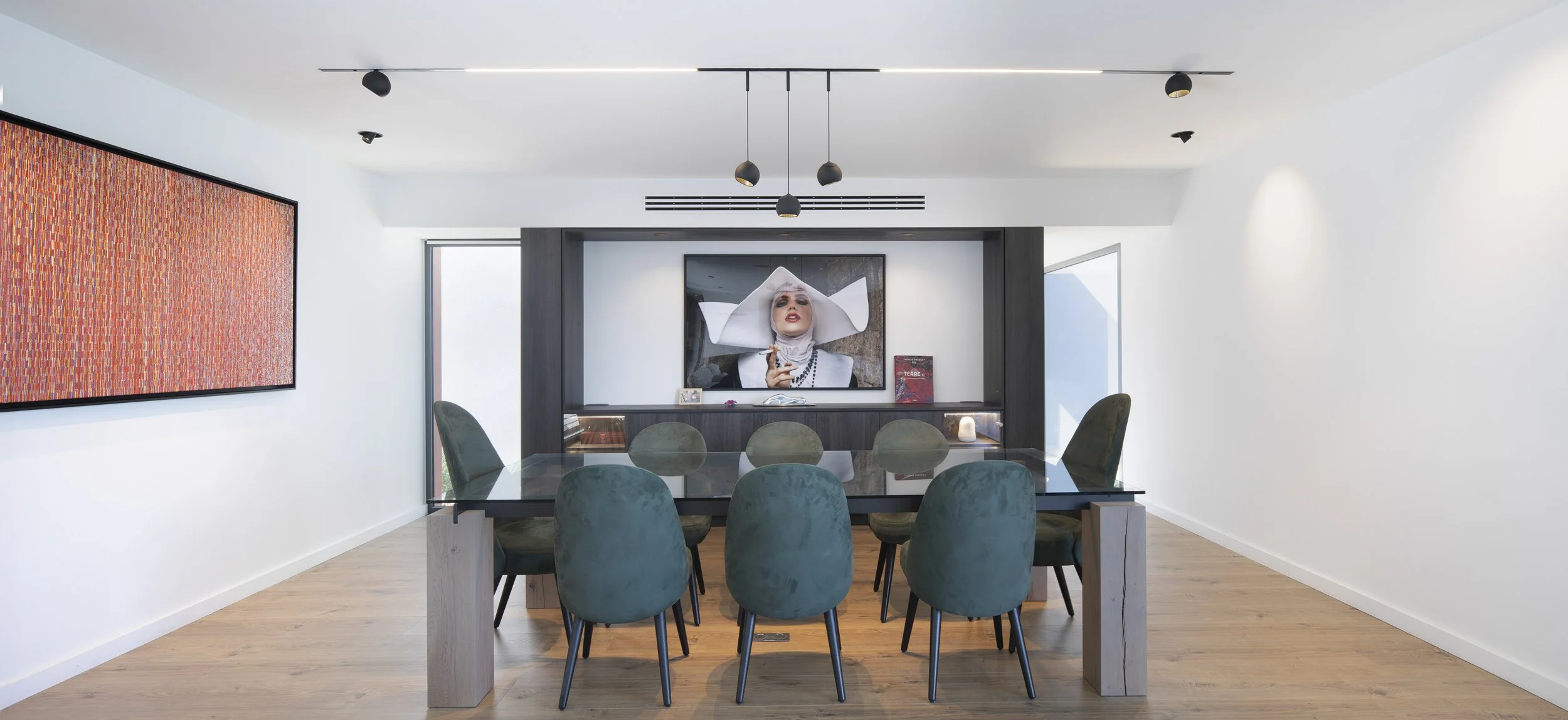 Salle à manger moderne avec huit chaises en velours vert autour d'une grande table en verre. Mur avec une grande peinture abstraite et une télévision affichant un portrait stylisé d'une femme portant un chapeau blanc. Sol en bois clair, murs blancs e