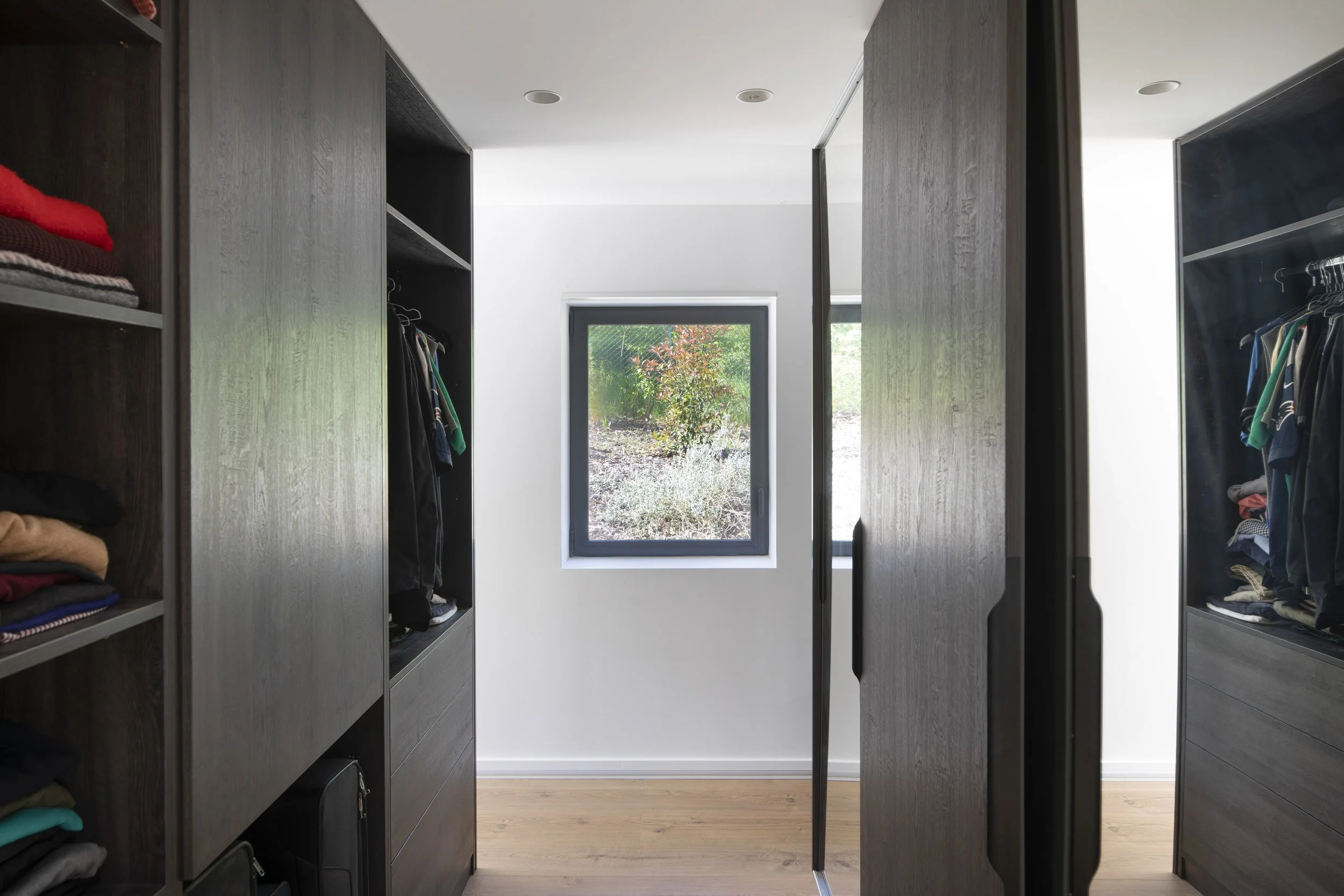 Un dressing avec des placards en bois foncé, ouverts, contenant des vêtements pliés et suspendus. Un mur blanc avec une fenêtre donnant sur un jardin en arrière-plan.