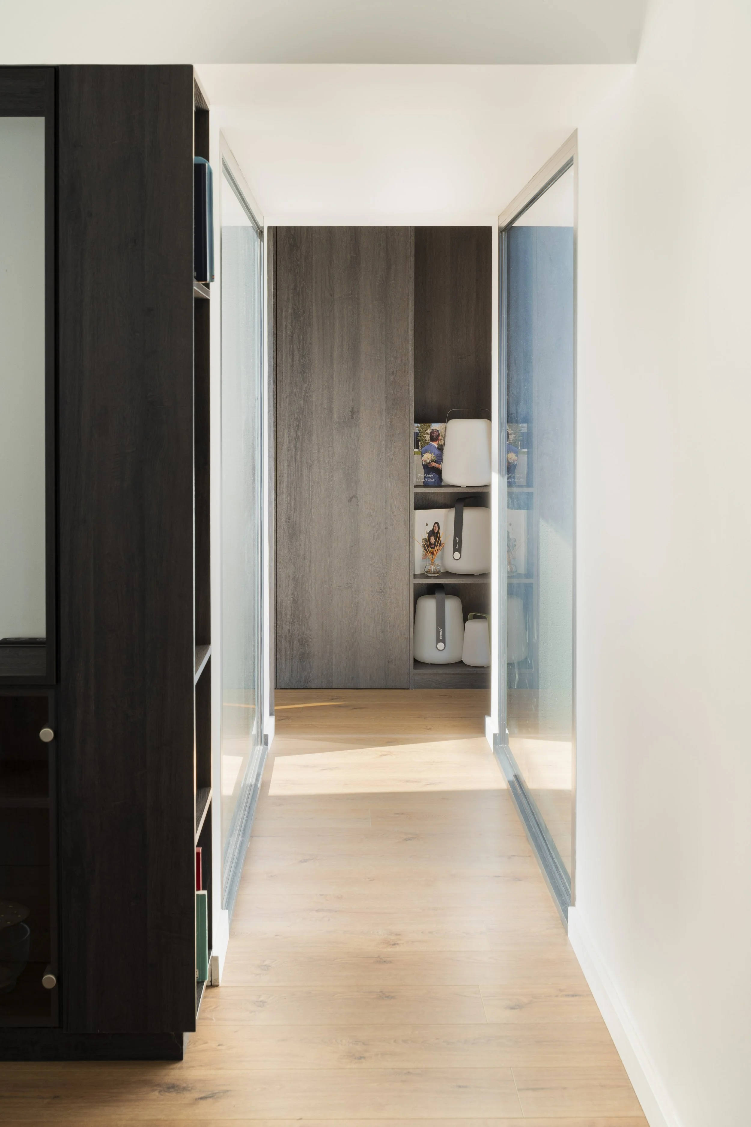Un couloir intérieur avec un sol en bois clair, murs blancs et un meuble en bois foncé avec des étagères contenant des objets blancs et un livre.