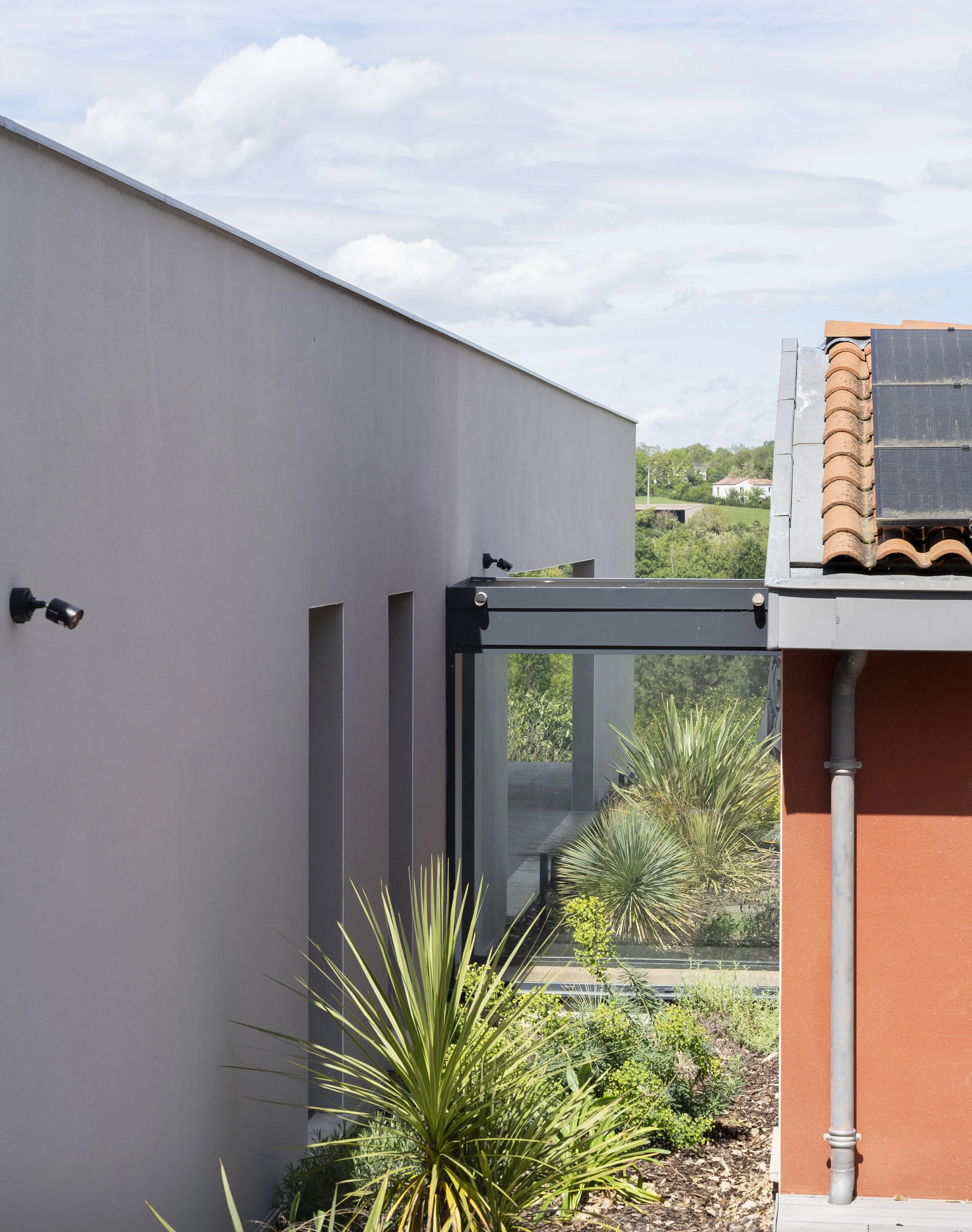 Vue d'une maison moderne avec mur gris, parterre de plantes tropicales et toiture en tuiles avec panneaux solaires.