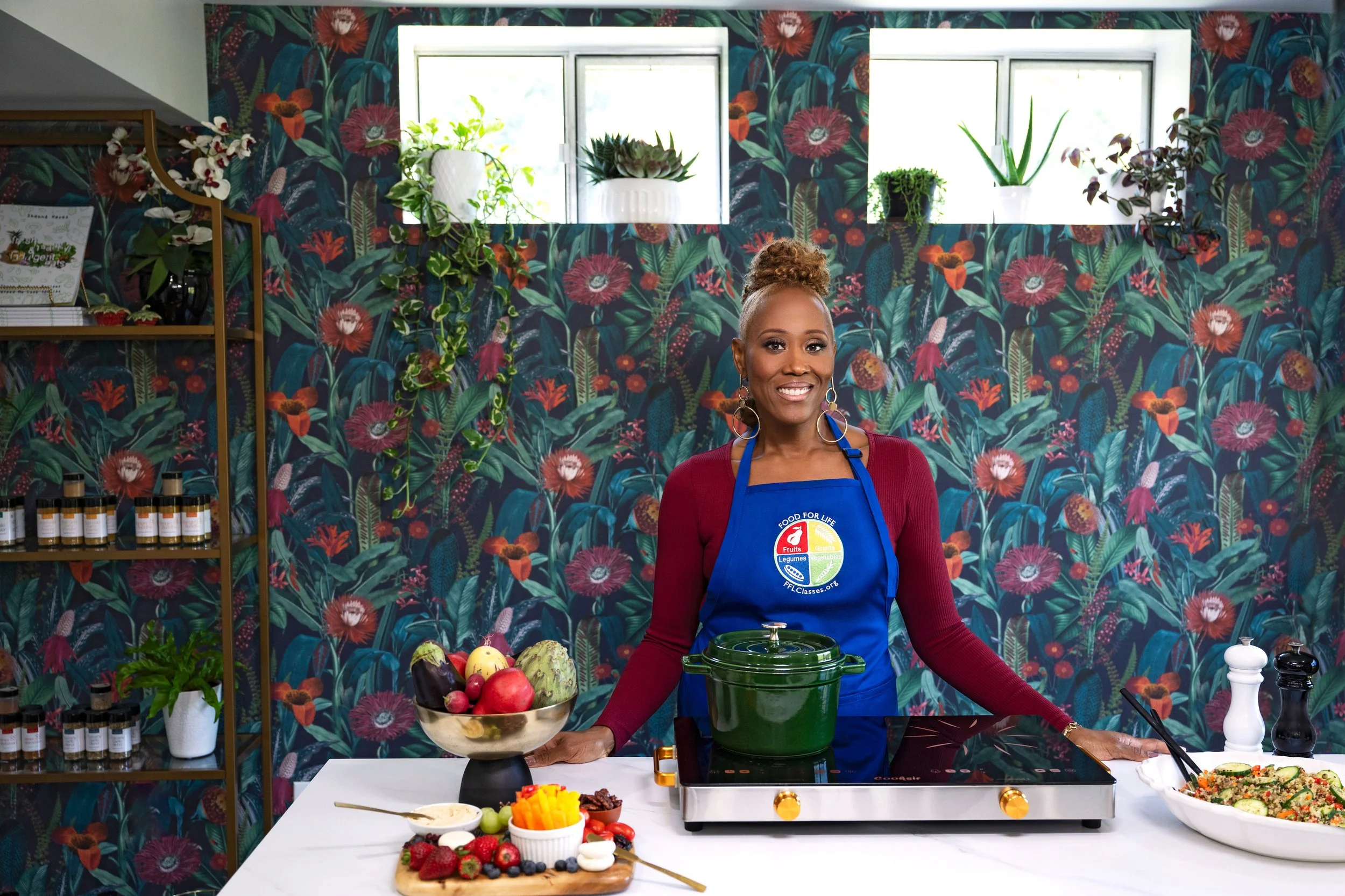 A smiling woman wearing a blue apron standing behind a kitchen counter with fresh fruits, vegetables, and a green pot, in a room with floral wallpaper and plants near the window.