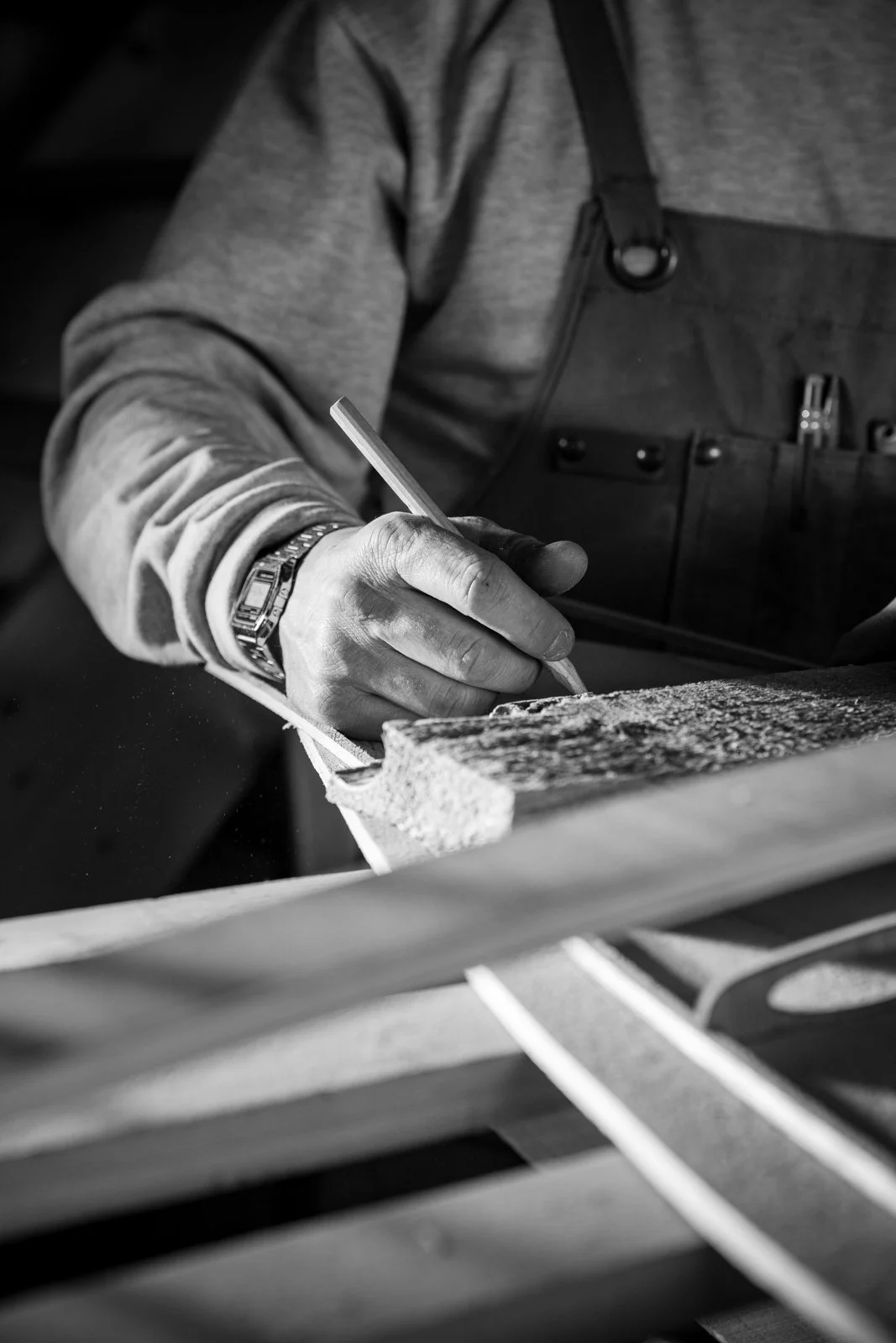 A person wearing a watch and a work apron is using a pencil to mark wood in a woodworking shop.