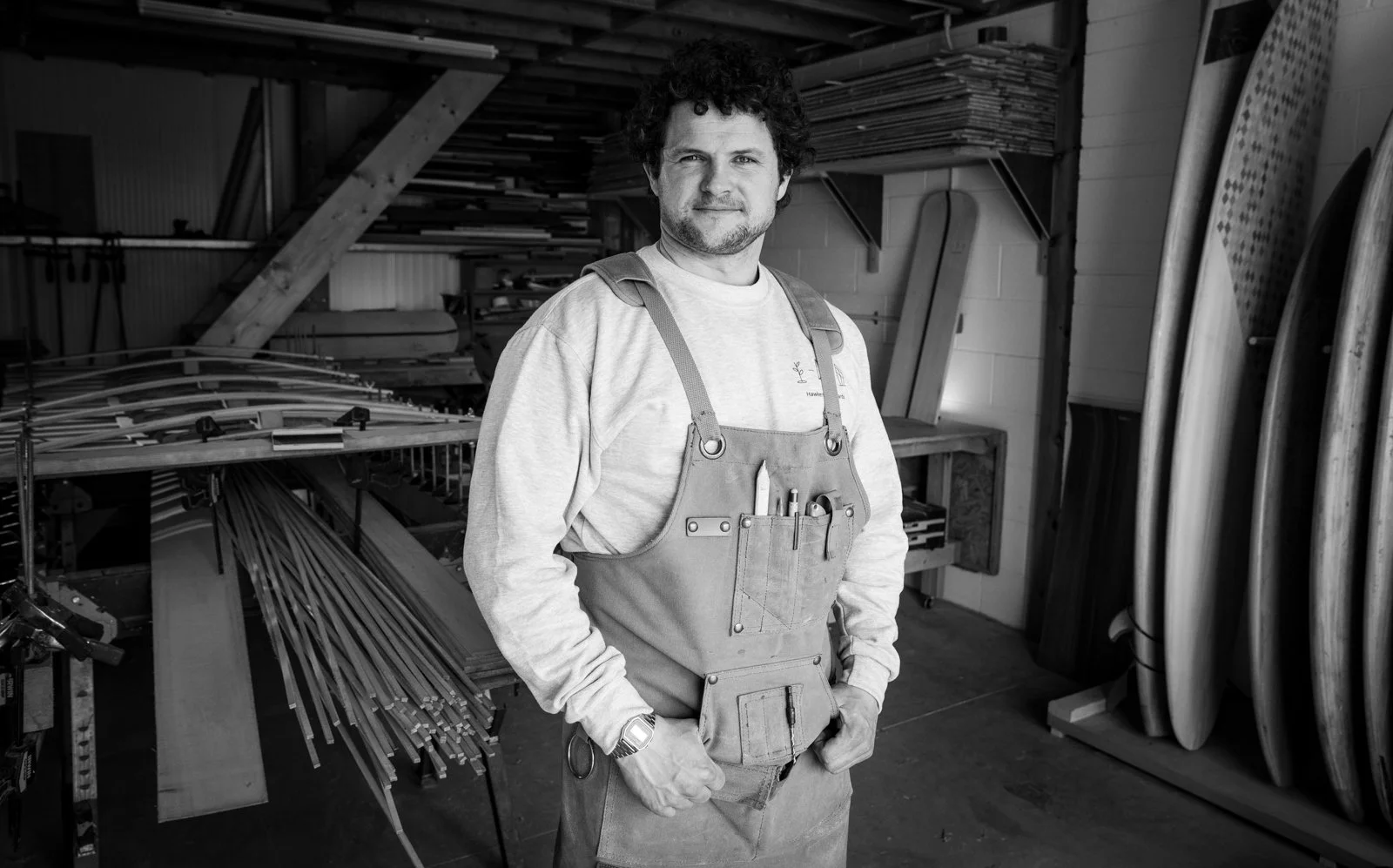A man with curly hair stands in a woodworking shop, wearing a work apron and posing with hands in pockets.