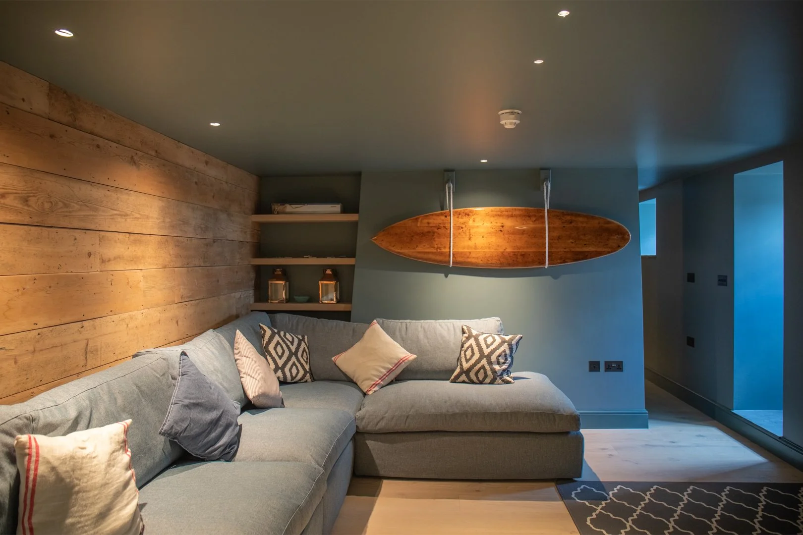 Living room with a light blue sectional sofa, multiple decorative pillows, a wooden surfboard mounted on the wall, a wooden wall to the left, and shelves with lanterns and decor. Part of a doorway and window are visible on the right.