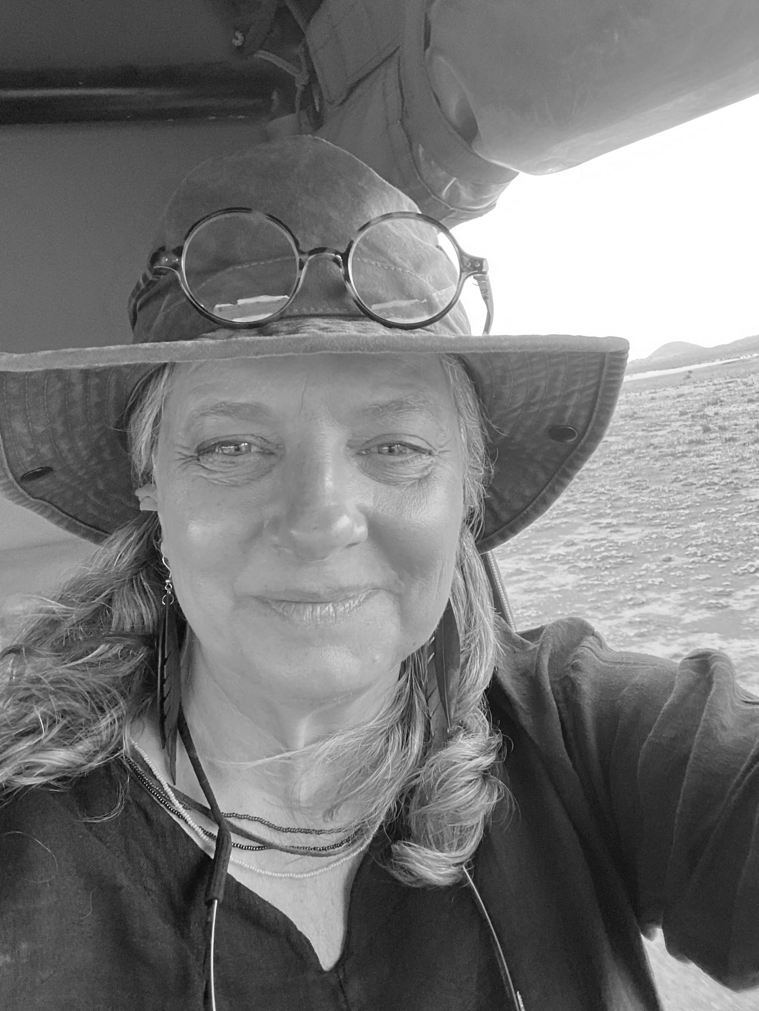 A woman with curly hair wearing a wide-brimmed hat, glasses on top of the hat, earrings, and a necklace, smiling outdoors with water and distant hills in the background, in black and white.