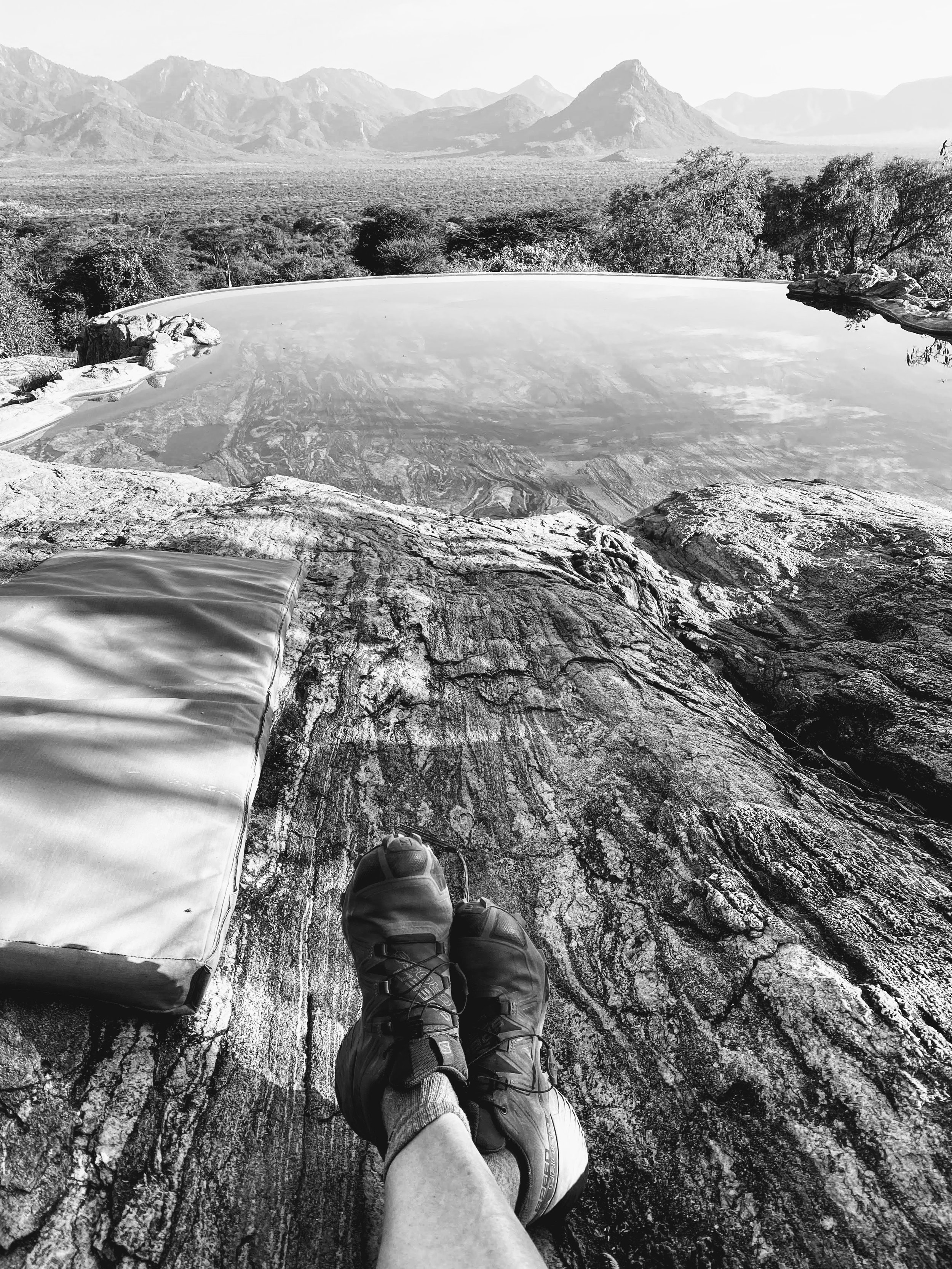 Person relaxing on a rock overlooking a natural hot spring with mountains in the background.