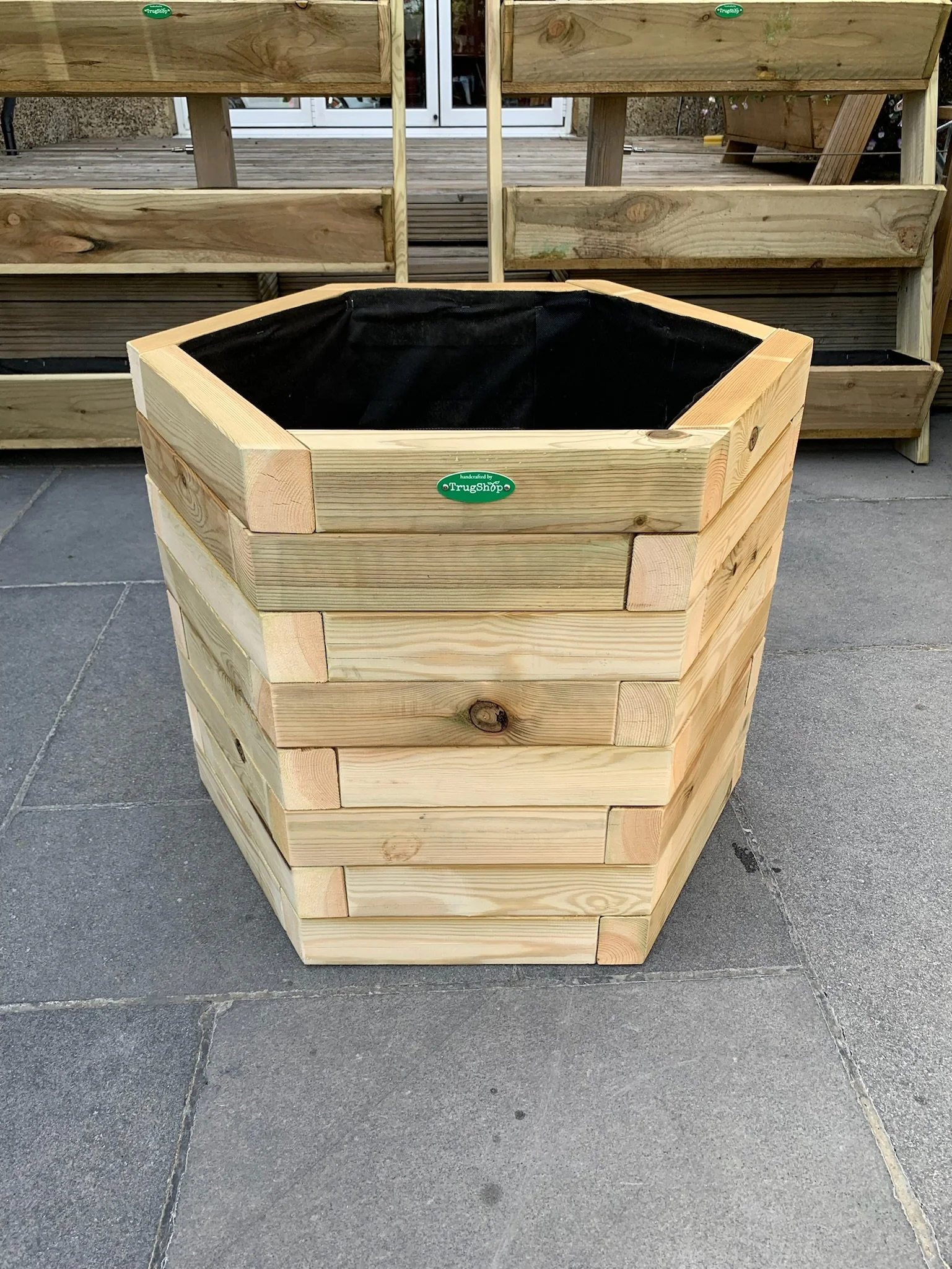 A large wooden hexagonal planter box with a black interior liner, placed on a tiled outdoor surface. In the background, there is a wooden deck and some gardening planters.