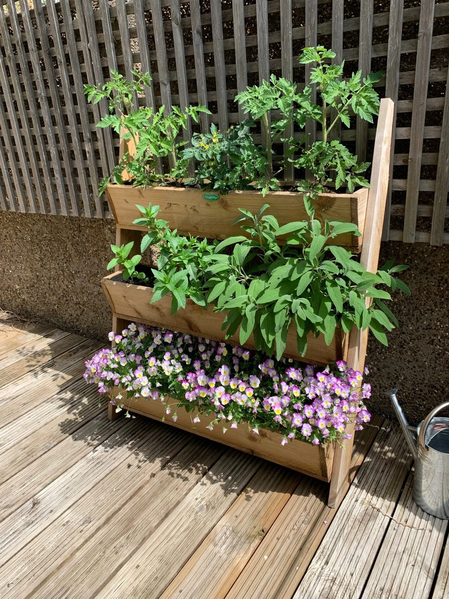 A tiered wooden planter with green leafy plants and blooming pink and white flowers on a wooden deck, with a lattice fence in the background and a watering can nearby.