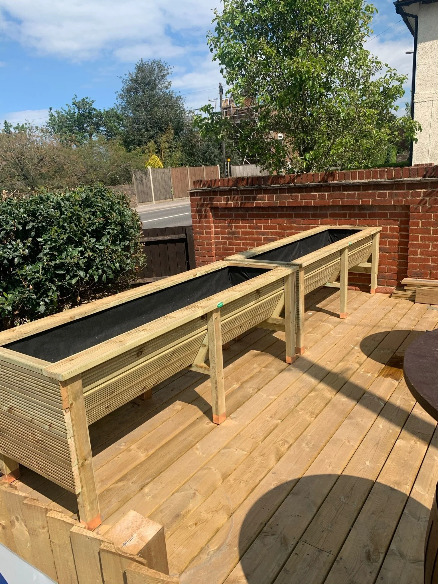 Three wooden planter boxes on a wooden deck with a brick wall, trees, and a street in the background.