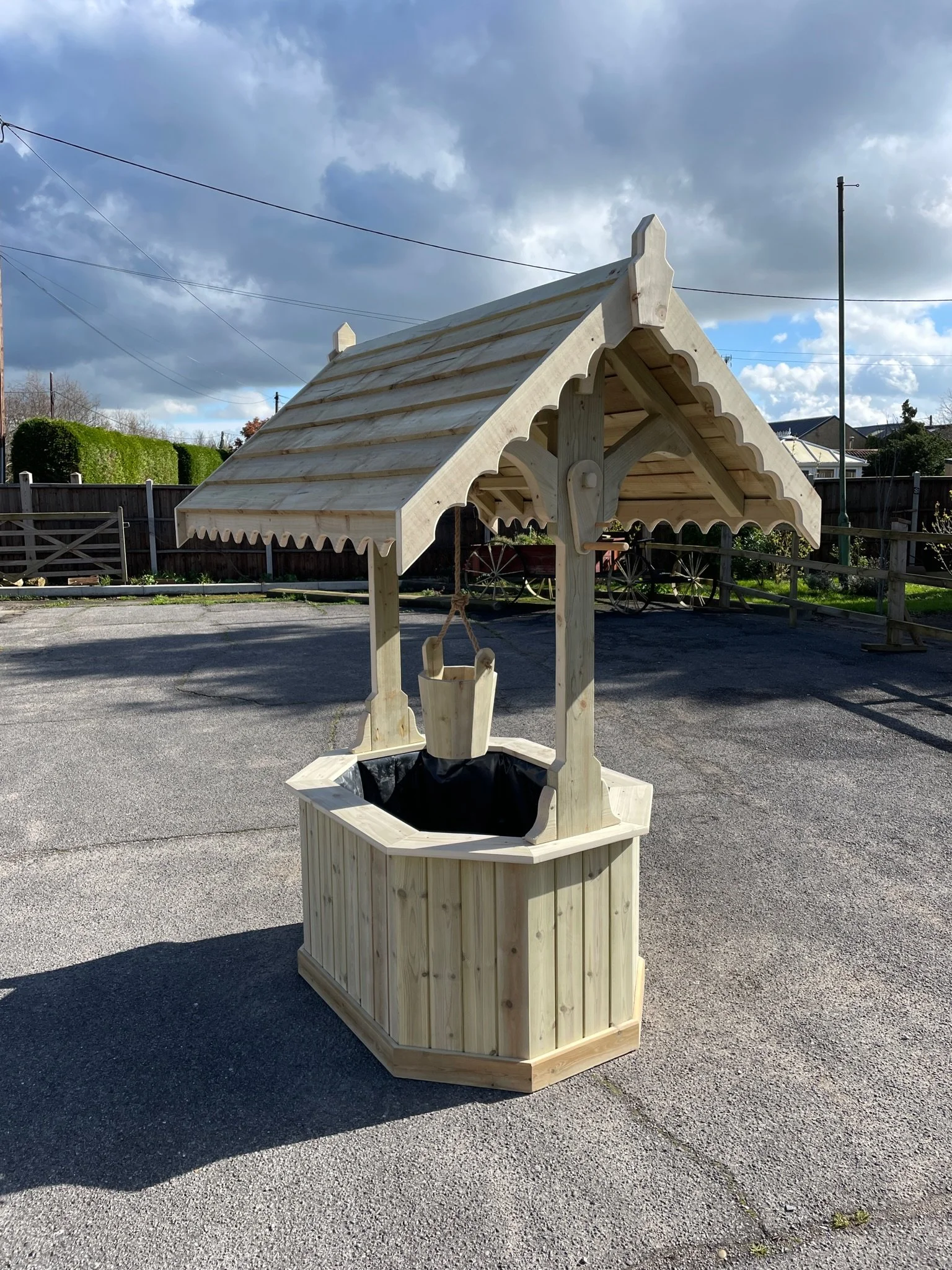 Empty wooden wishing well with a roof and a bucket hanging from a rope, situated outdoors on a paved surface.