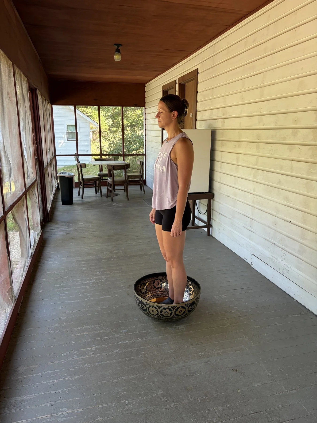 A woman standing inside a porch with her feet submerged in a singing bowl, eyes closed, wearing a sleeveless top and shorts, with trees and a white house visible outside. Sara Taylor Wellness Experience