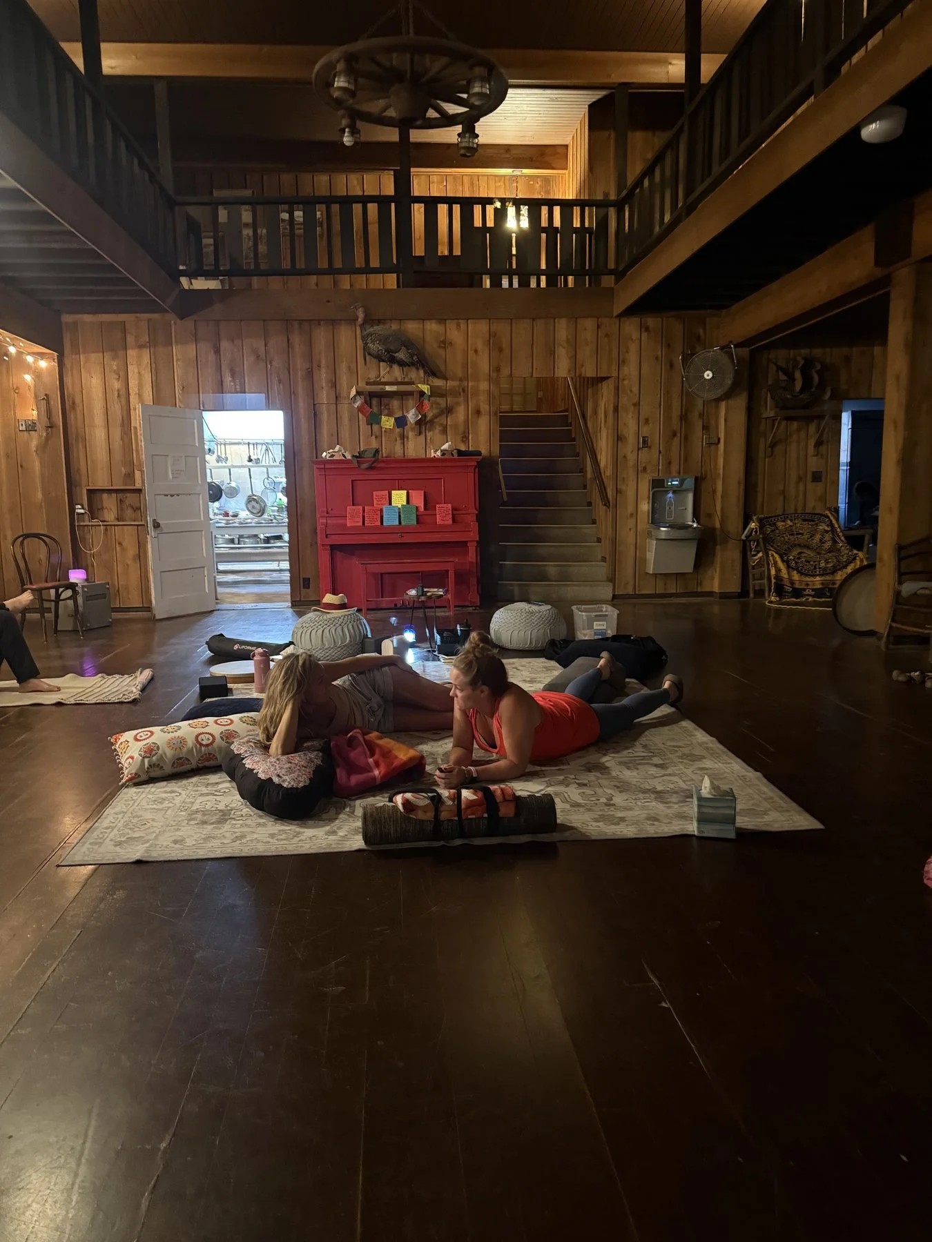 The image shows an indoor space with wooden walls and a high ceiling, where several people are relaxing on a rug on the wooden floor. Some are lying down, and others are sitting and chatting. The room has cozy decor, including a mounted turkey, a red