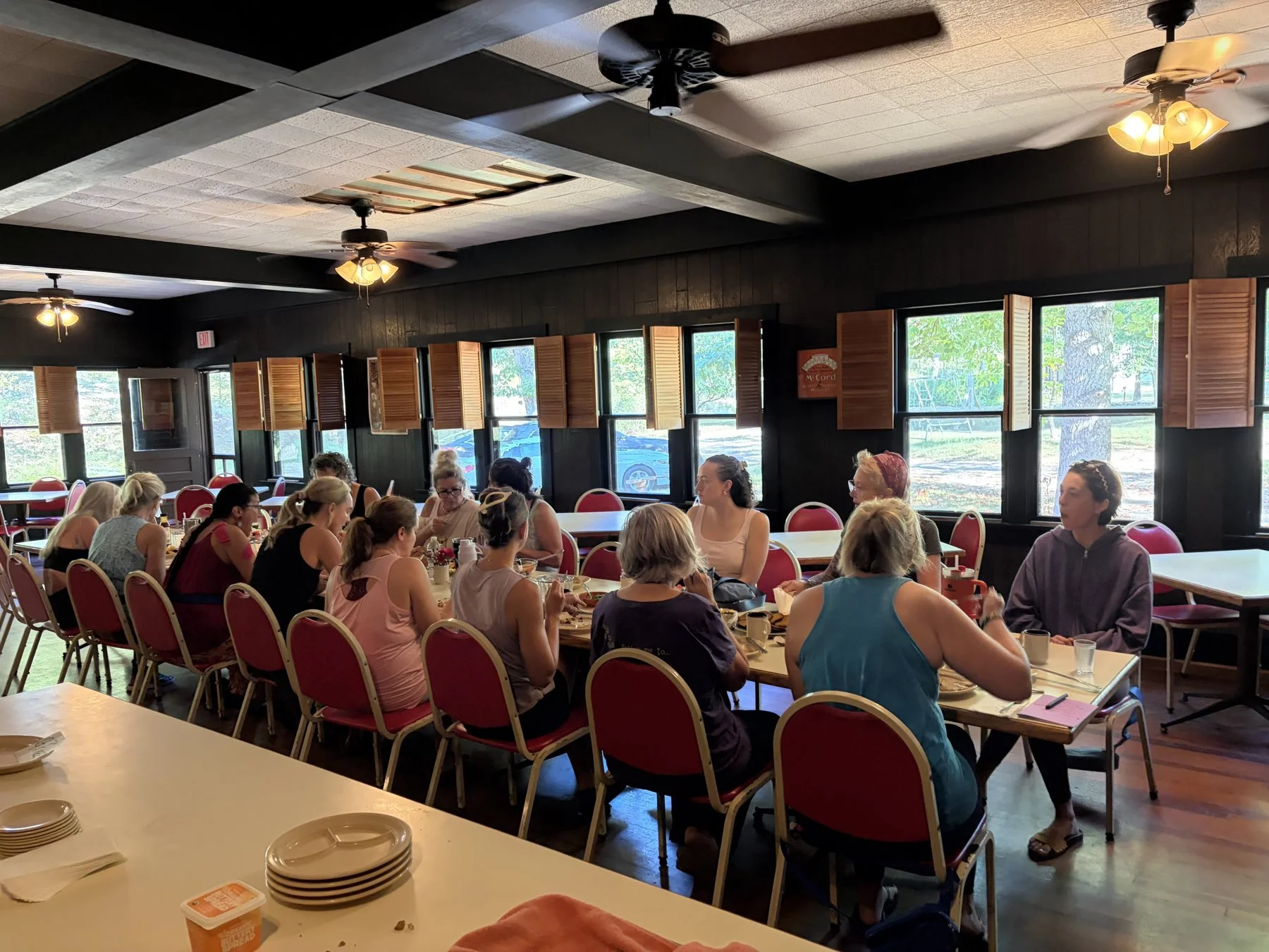 A group of women sitting at long tables in a dining hall, engaged in conversation and eating. The room has large windows with open wooden shutters, black walls, a wooden floor, and ceiling fans with lighting. The scene appears casual and social.