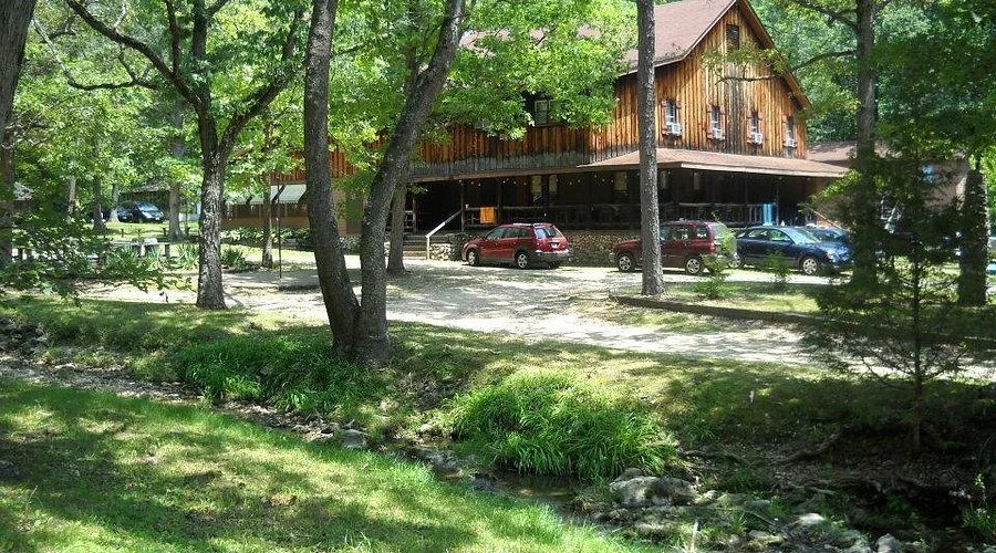 A rustic wooden building surrounded by green trees with parked cars in front, near a small creek in a grassy area.