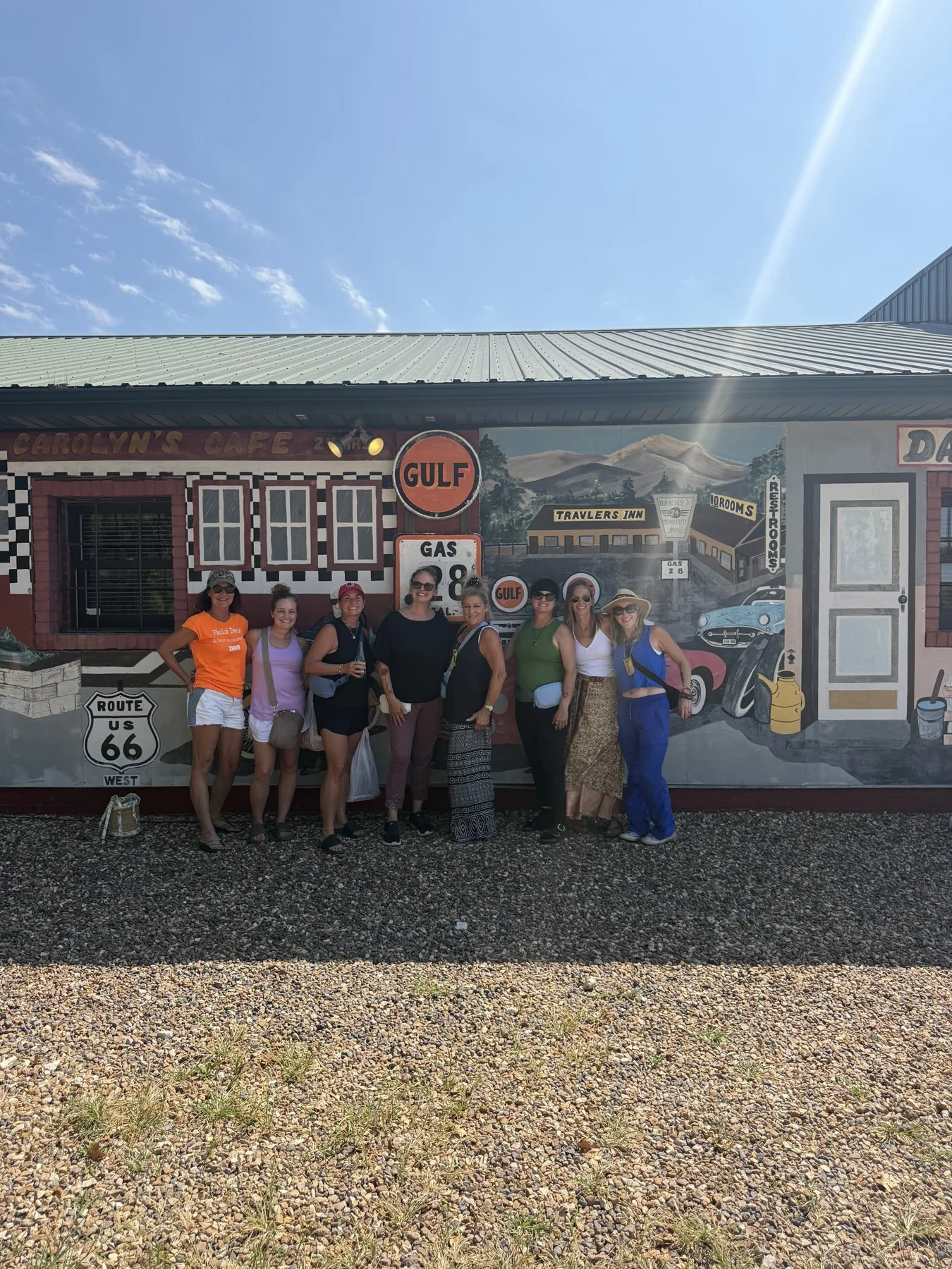 A group of eight women standing in front of a mural on a building, with a clear blue sky in the background. Sara Taylor Wellness Experience