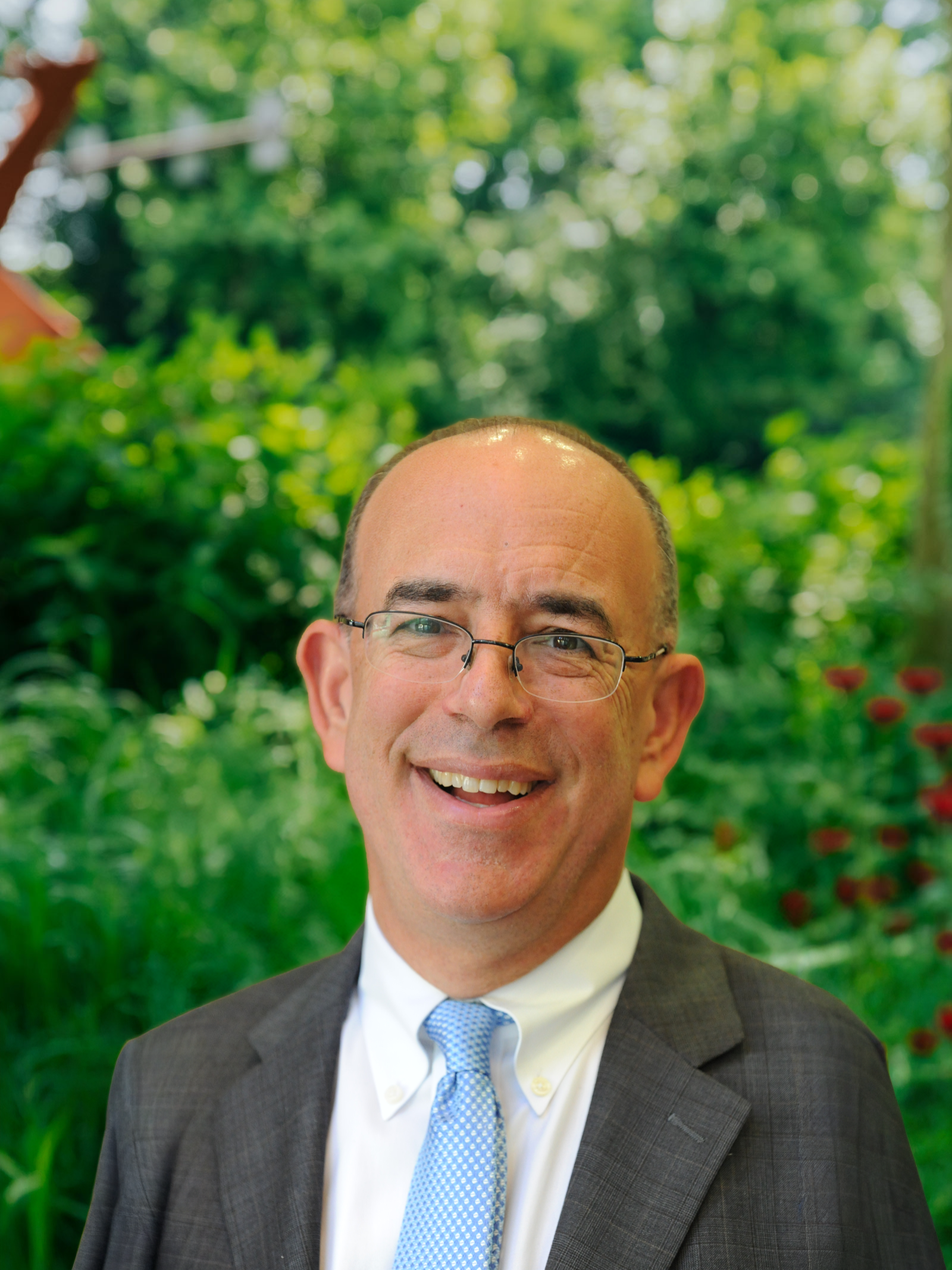 Smiling man wearing glasses, a suit, and a blue tie outdoors with green foliage background.