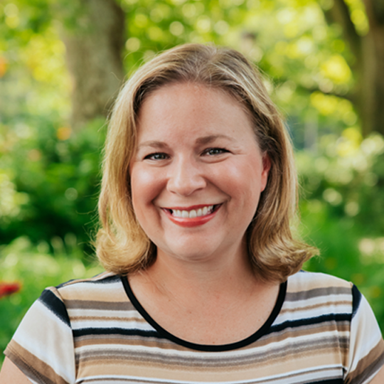 Smiling woman with shoulder-length blonde hair outdoors, wearing a striped T-shirt, with greenery and trees in the background.