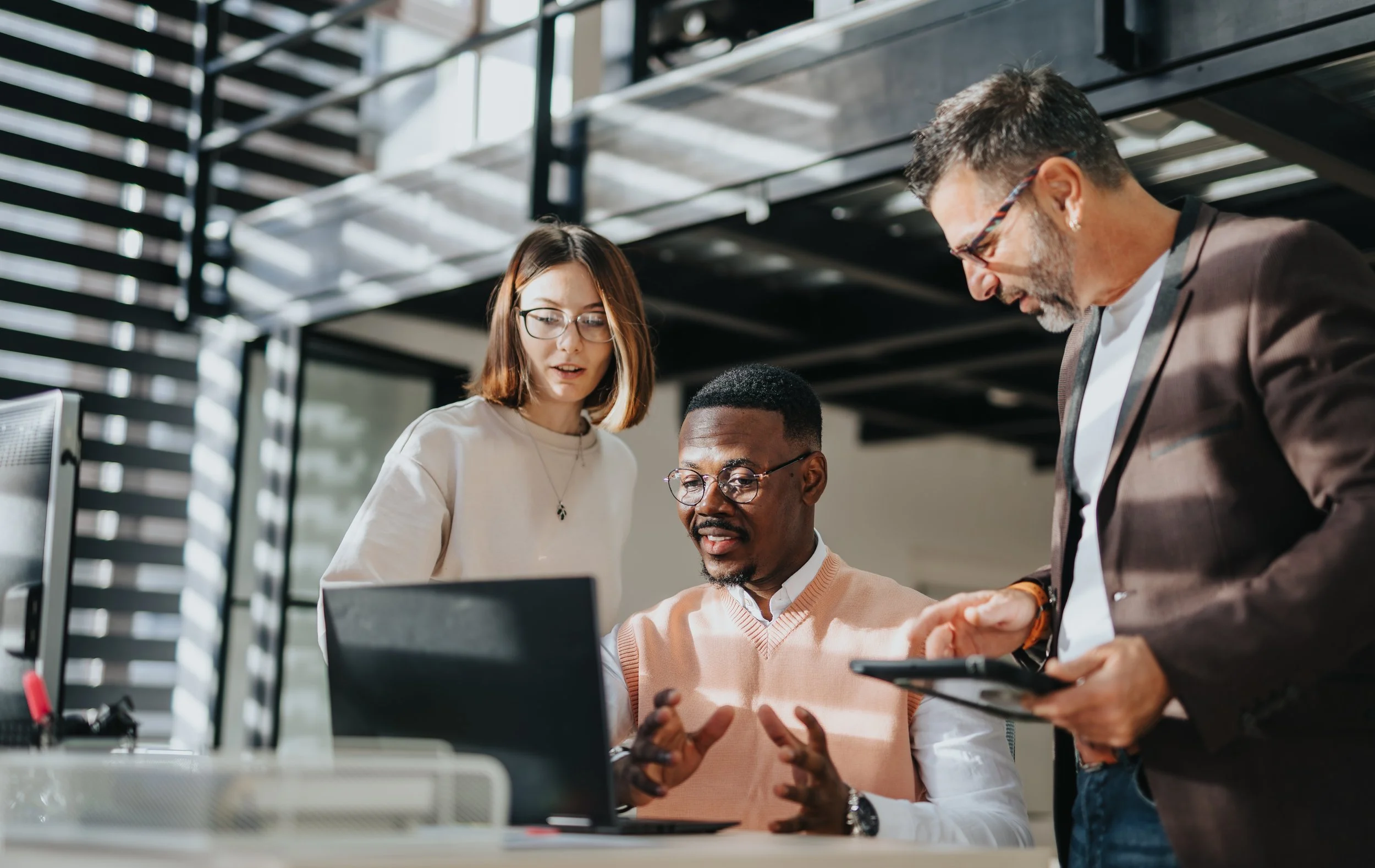 Three diverse professionals in a modern office discussing work around a laptop and a tablet.