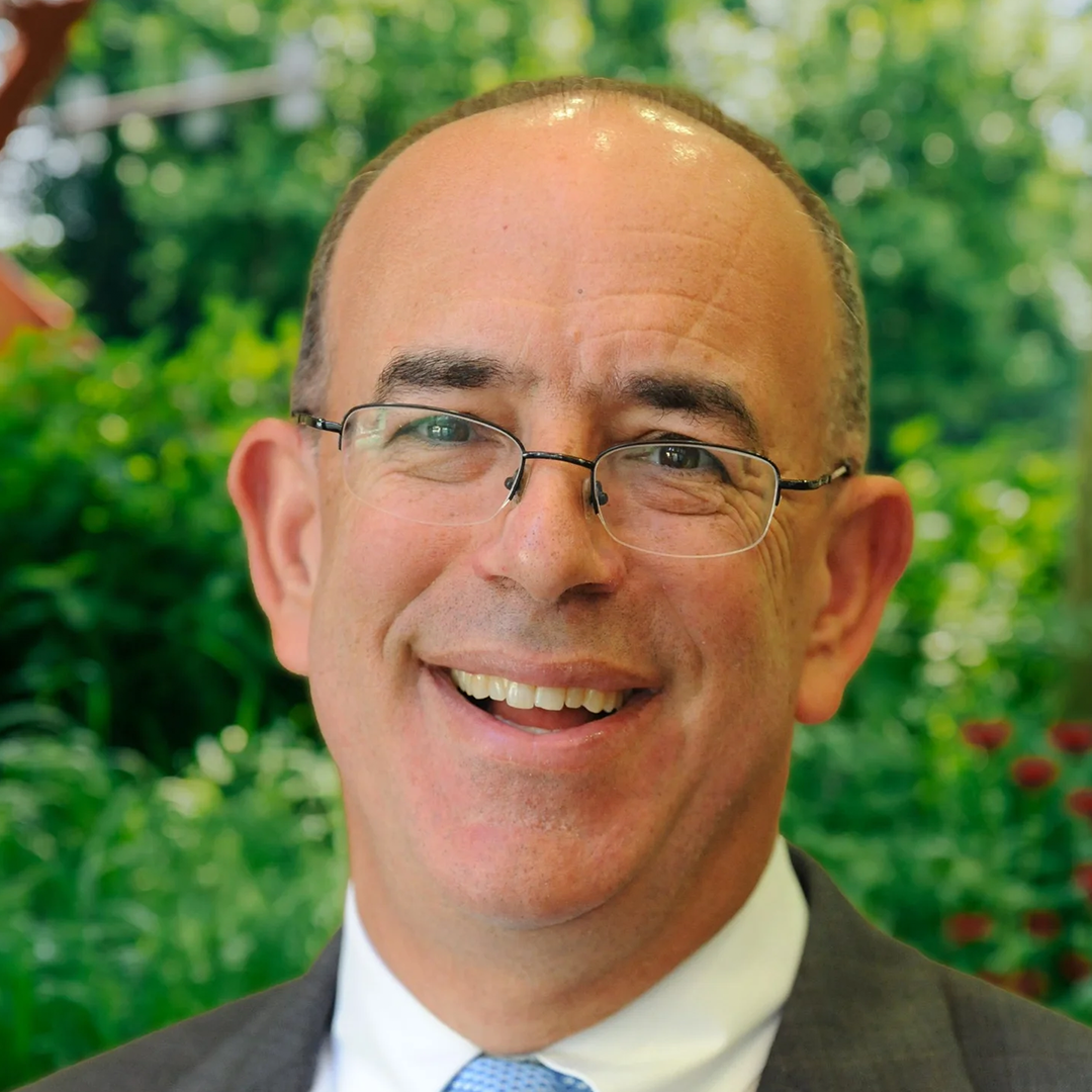 Smiling man with glasses in suit and tie outdoors amid green foliage.