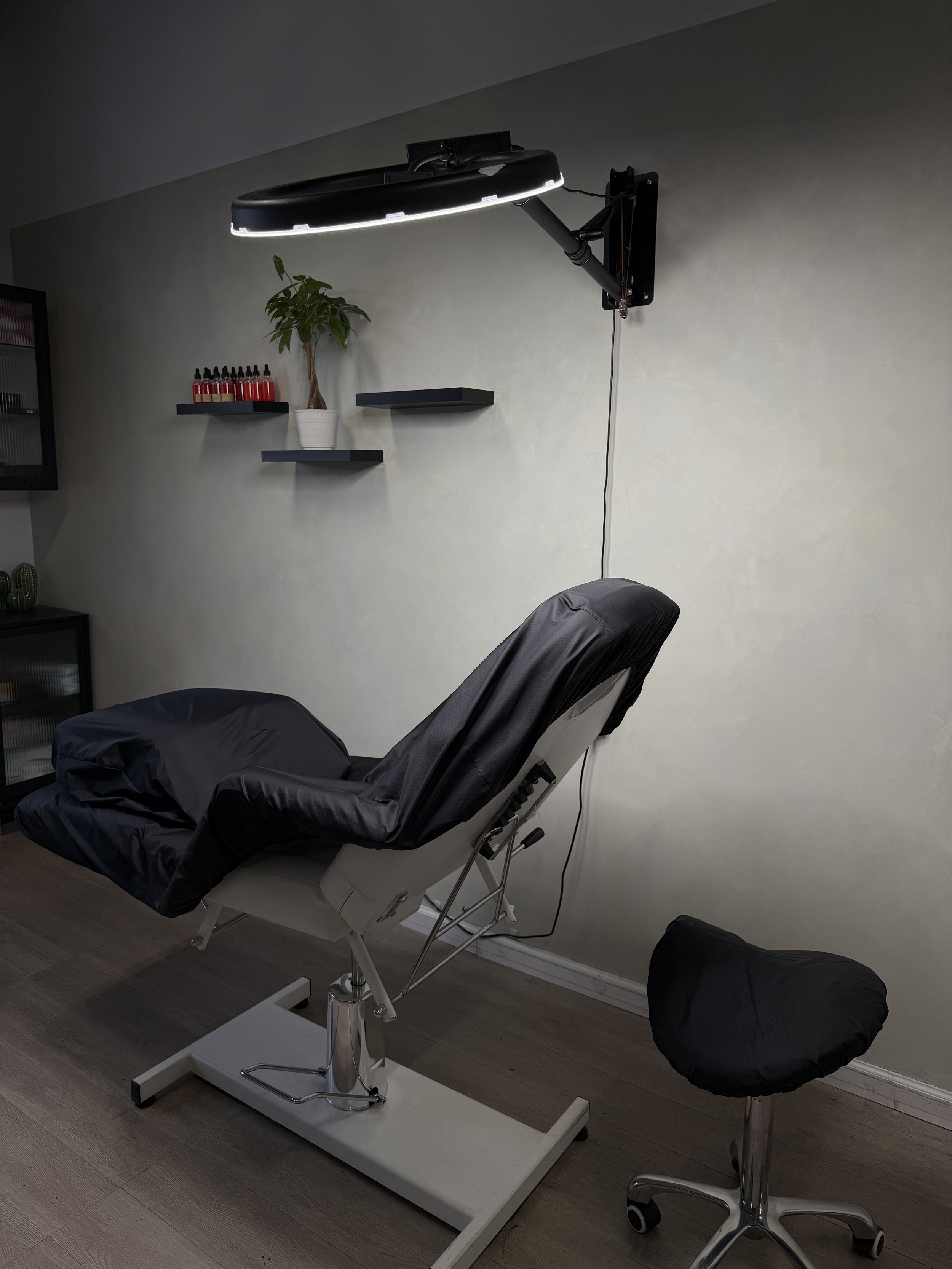 A modern medical or massage treatment room with an adjustable black examination chair, a black chair with a matching footrest, a wall-mounted light with a circular design, a potted plant, and small bottles on shelves, with wood flooring.