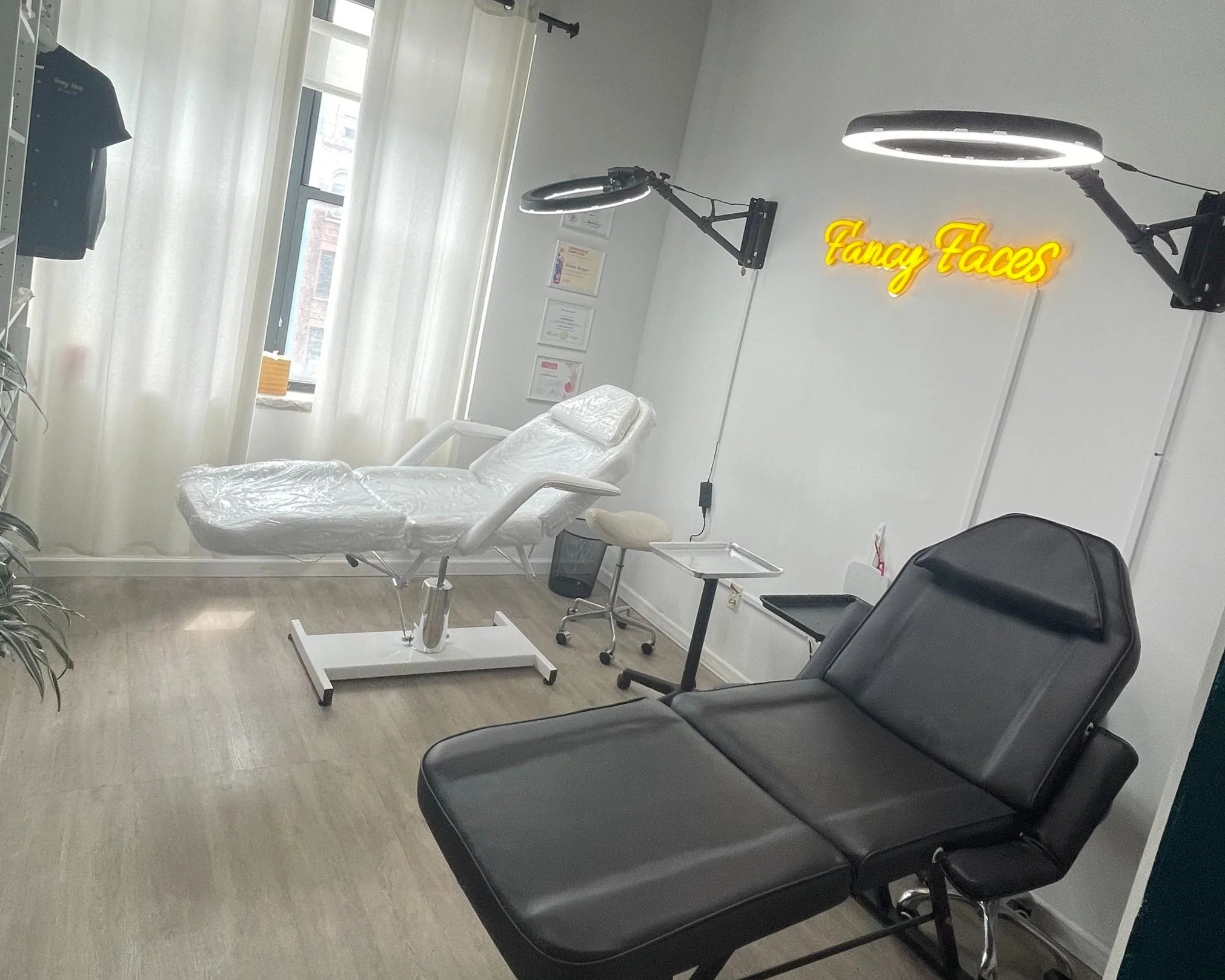 Medical examination room with a black reclining chair, a white examination bed with plastic cover, drawing lights overhead, a small rolling stool, and a neon sign on the wall that says "Fancy Faces". There are framed certificates on the wall, a window with white curtains, and some plants.