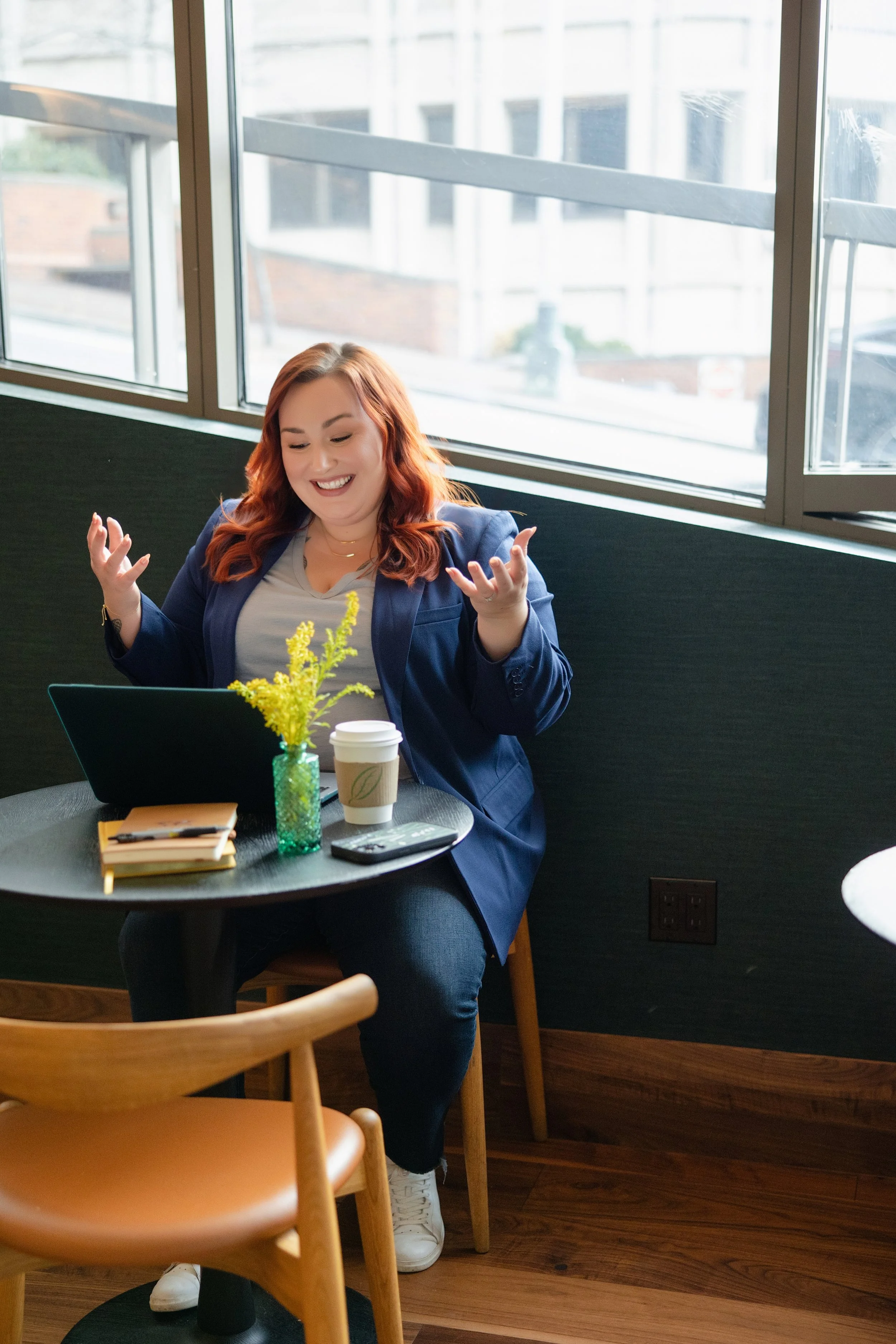 A woman sitting at a small round table in a cafe, smiling and gesturing with her hands. The table holds a small vase with yellow flowers, a virtual meeting device, a coffee cup, a notebook, and a smartphone. Large windows behind her let in natural light.