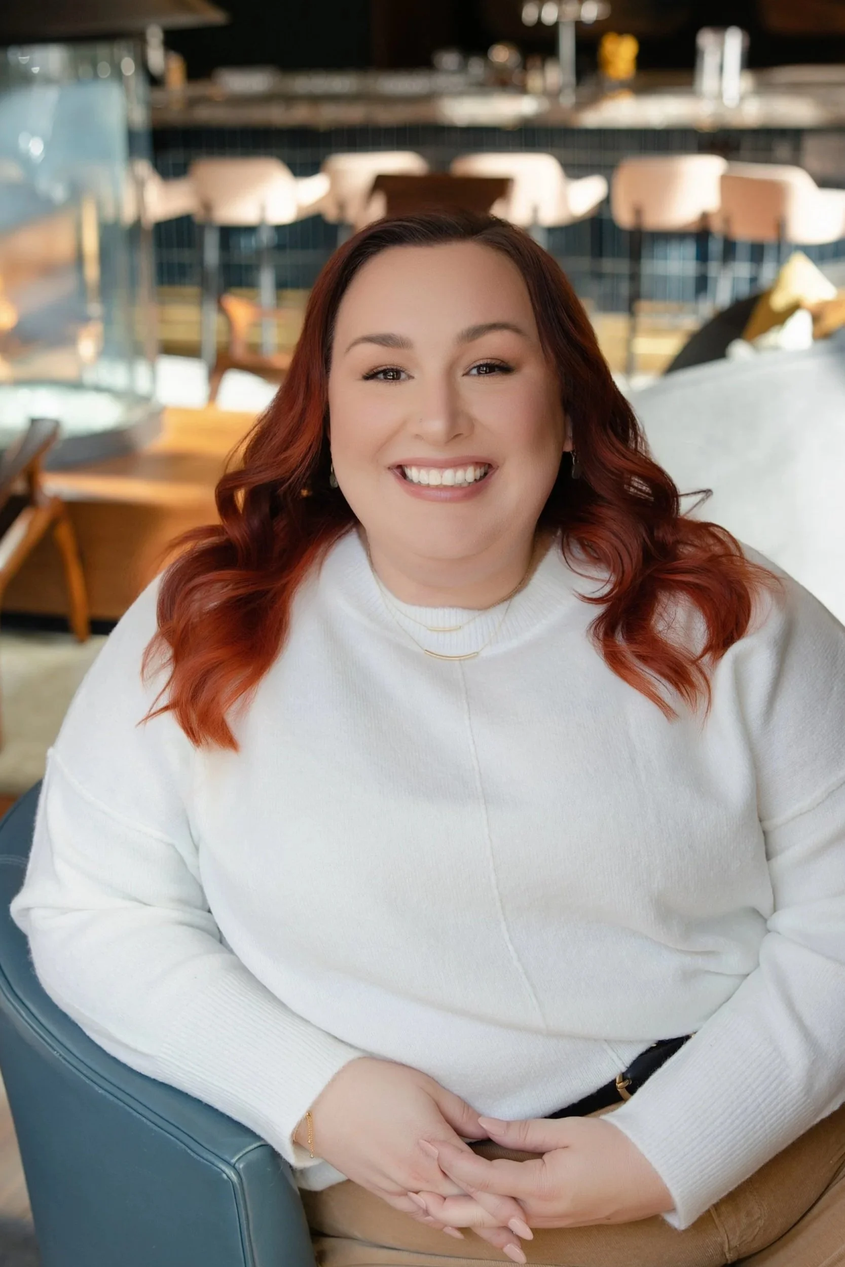 A woman with red hair smiling, seated indoors in a restaurant or cafe setting.