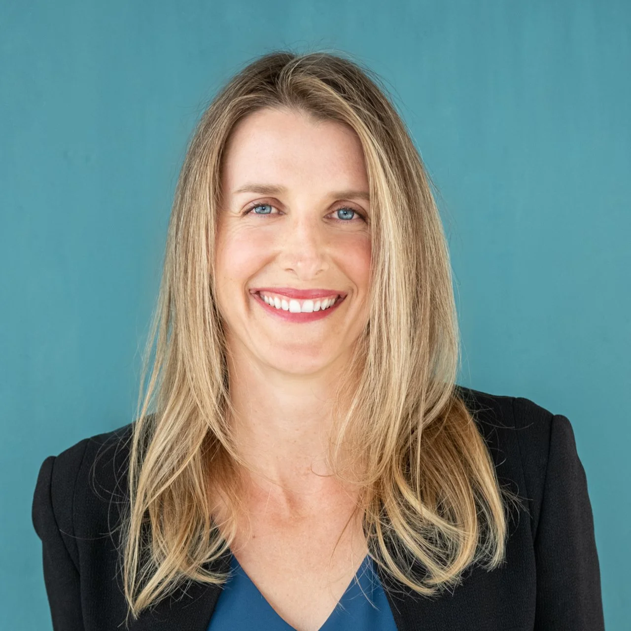 A woman with blonde hair, blue eyes, and a big smile, wearing a black blazer over a blue top, standing against a teal background.
