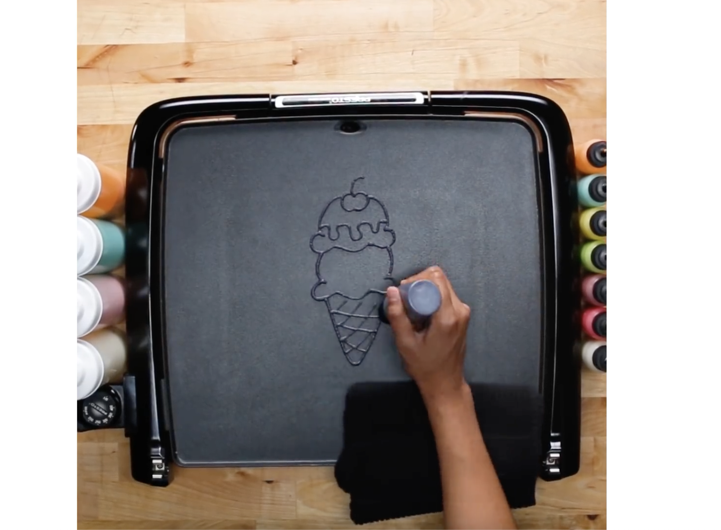 Hand drawing an ice cream cone with whipped cream and a cherry on top on a black textured surface surrounded by bottles of colorful markers. Pancake art, Pancake art party, Pancake artist for hire, Pancake artist near me, Pancake artist Los Angeles