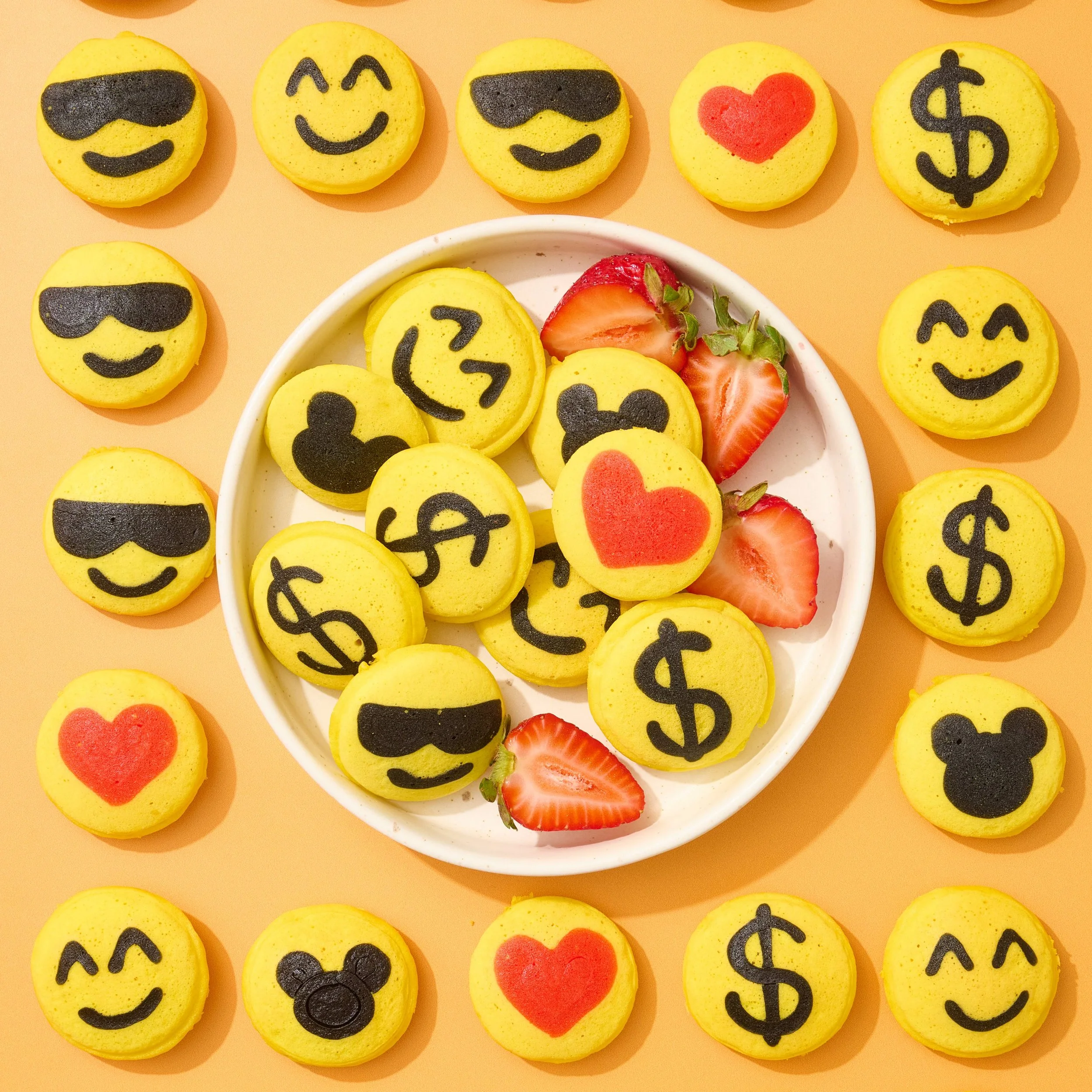 Yellow pancakes with black emojis such as heart eyes, sunglasses, smiling face, dollar sign, and Mickey Mouse, arranged around a white bowl containing similar cookies and fresh strawberries.  Pancake art, Pancake art party, Pancake artist for hire