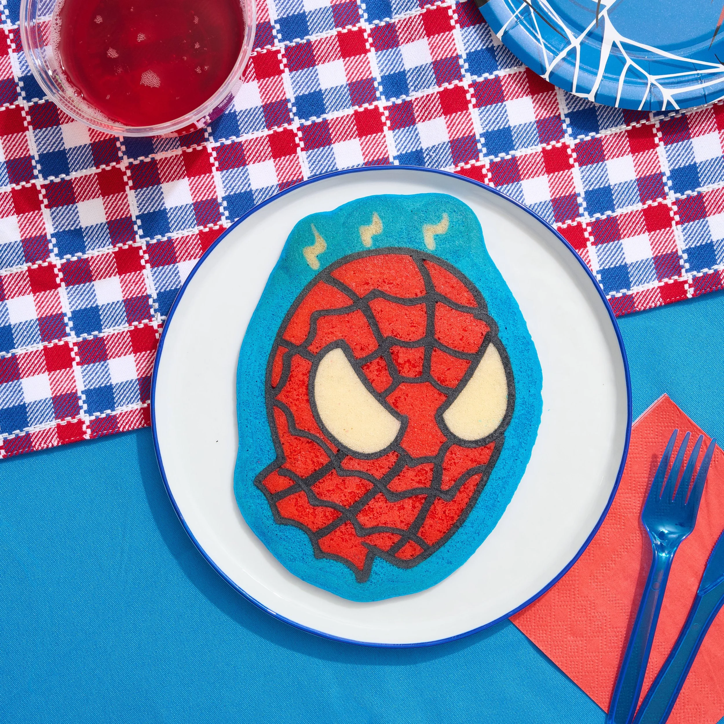 A  pancake shaped like Spider-Man's mask on a white plate with a blue rim, placed on a blue table with a red, white, and blue checkered tablecloth, and a red napkin with a blue fork and knife next to it. Pancake art, Pancake art party, Pancake artist
