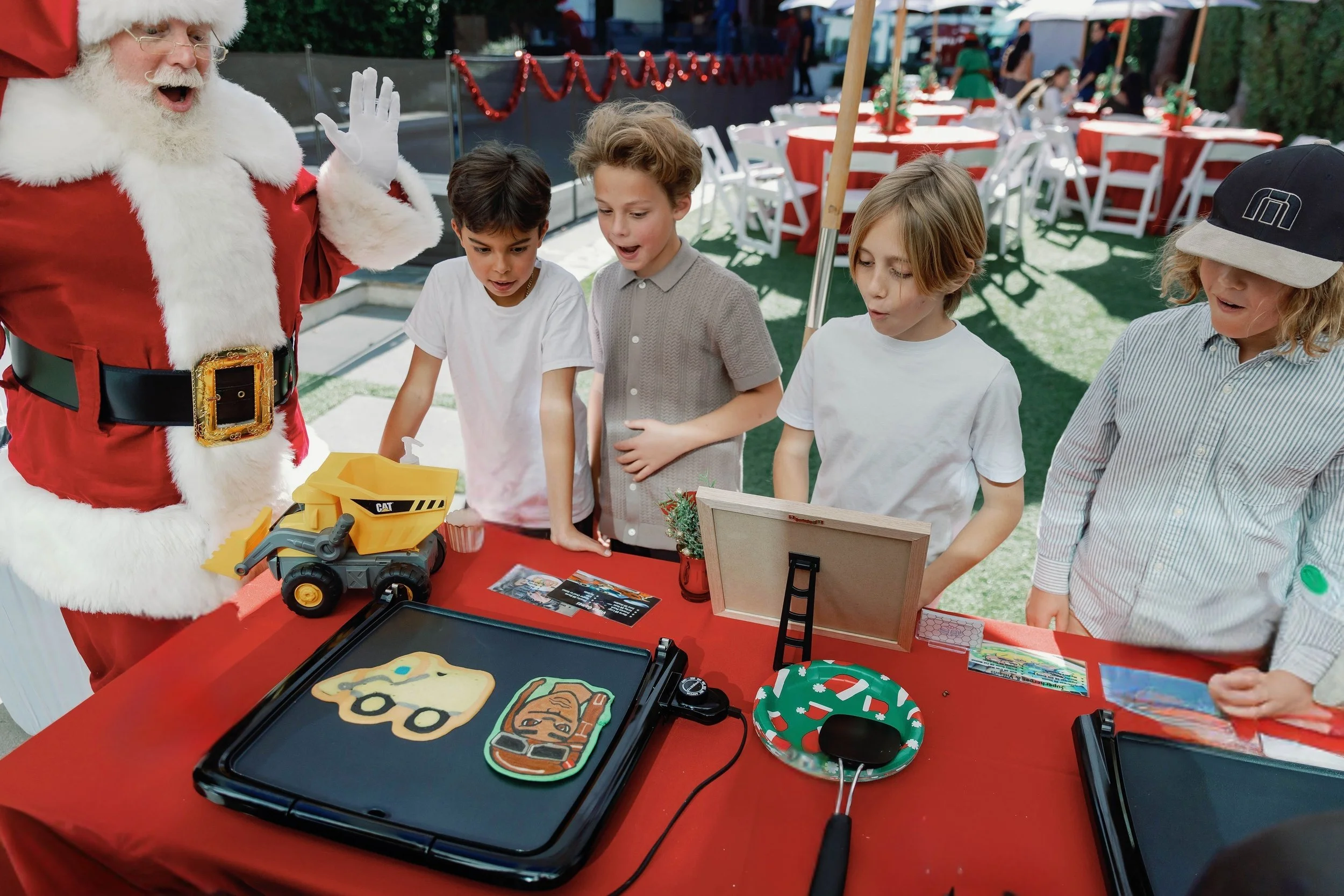 Santa Claus in a red suit and black belt with gold buckle talking to children at a holiday event, with decorated tables and children in the background.  Pancake art, Pancake art party, Pancake artist for hire, Pancake artist near me, Pancake artist