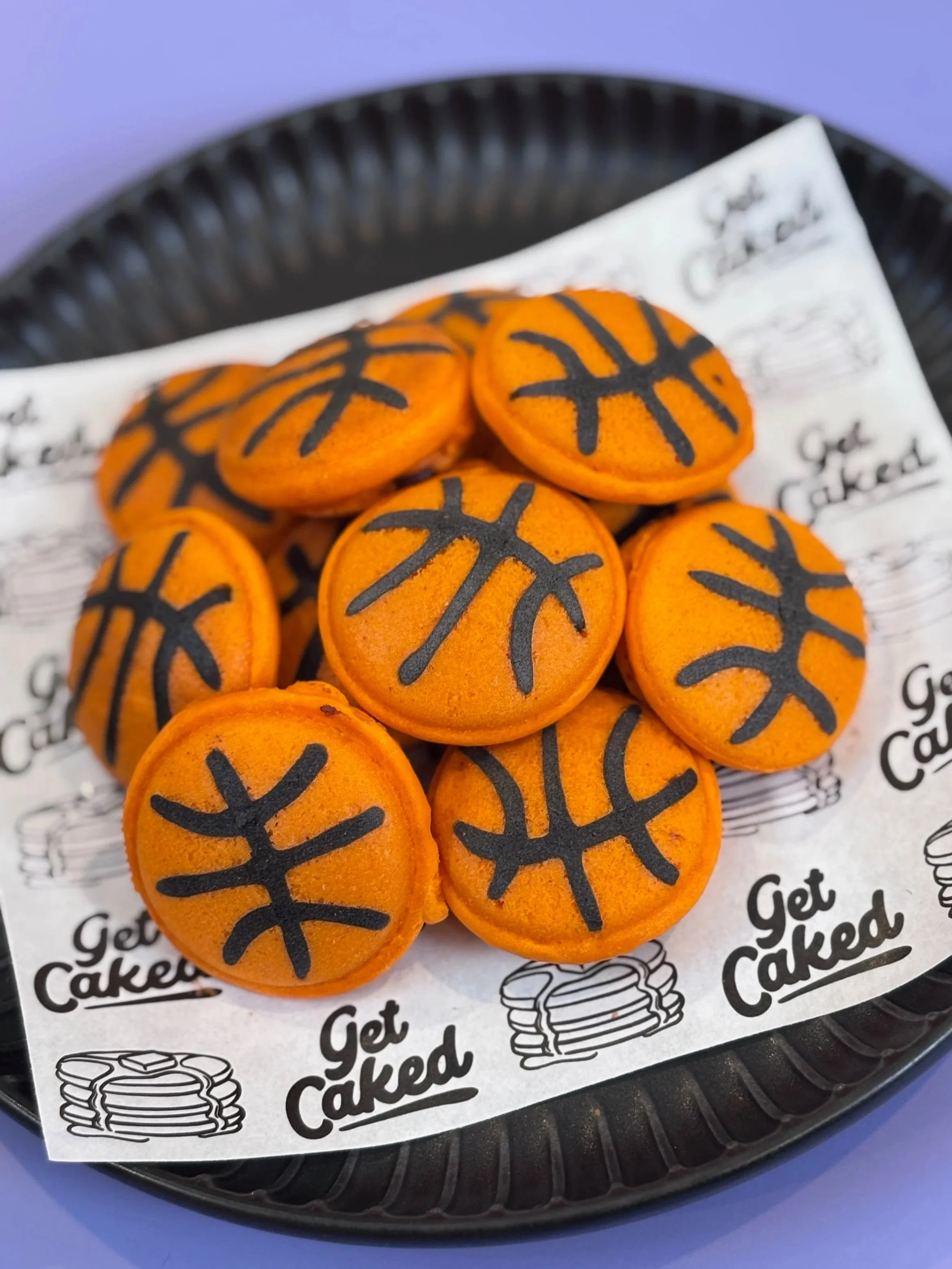 Orange football-shaped cookies with black laces design on top, placed on a white paper with 'Get Caked' printed on it, on a black tray. Mini pancake art,  Pancake art, Pancake art party, Pancake artist for hire, Pancake artist near me