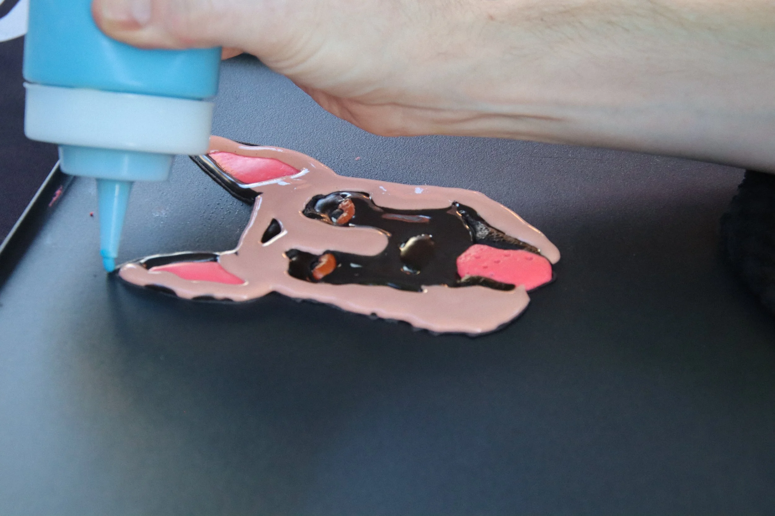 A person uses a squeeze bottle to decorate a cake with pink, black, and brown icing in the shape of a dog face.  Pancake art, Pancake art party, Pancake artist for hire, Pancake artist near me