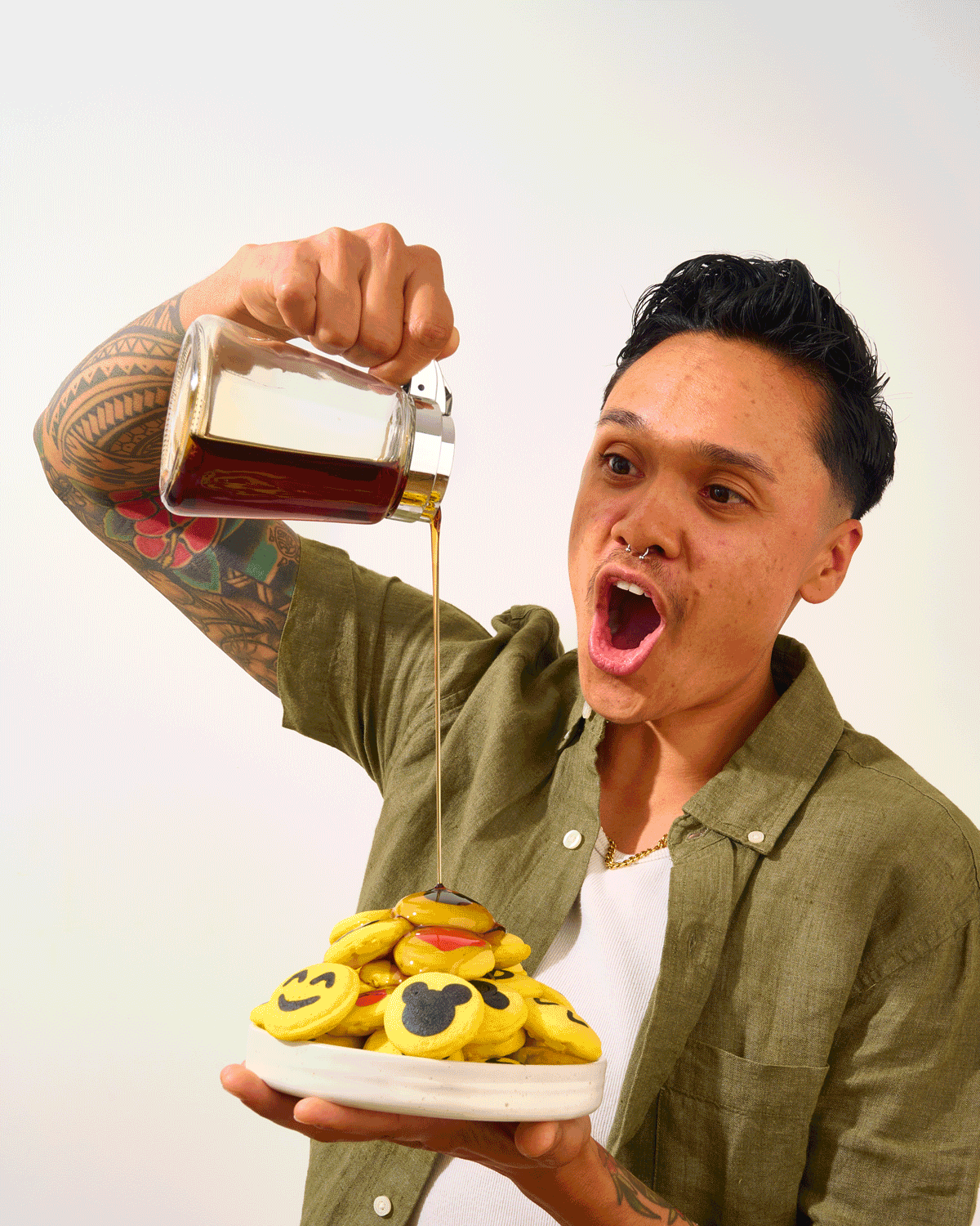 A person with tattoos and a septum piercing pouring syrup on a plate of emoji-shaped cookies, with an excited facial expression.  Pancake art, Pancake art party, Pancake artist for hire, Pancake artist near me