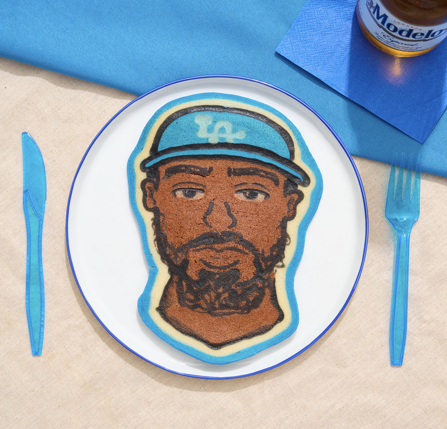 Cookie decorated with a portrait of a man wearing a hat with a dollar sign, on a white plate, with blue fork and knife, beige tablecloth, blue napkin, and a bottle of Modelo beer in the background. LA Dodgers