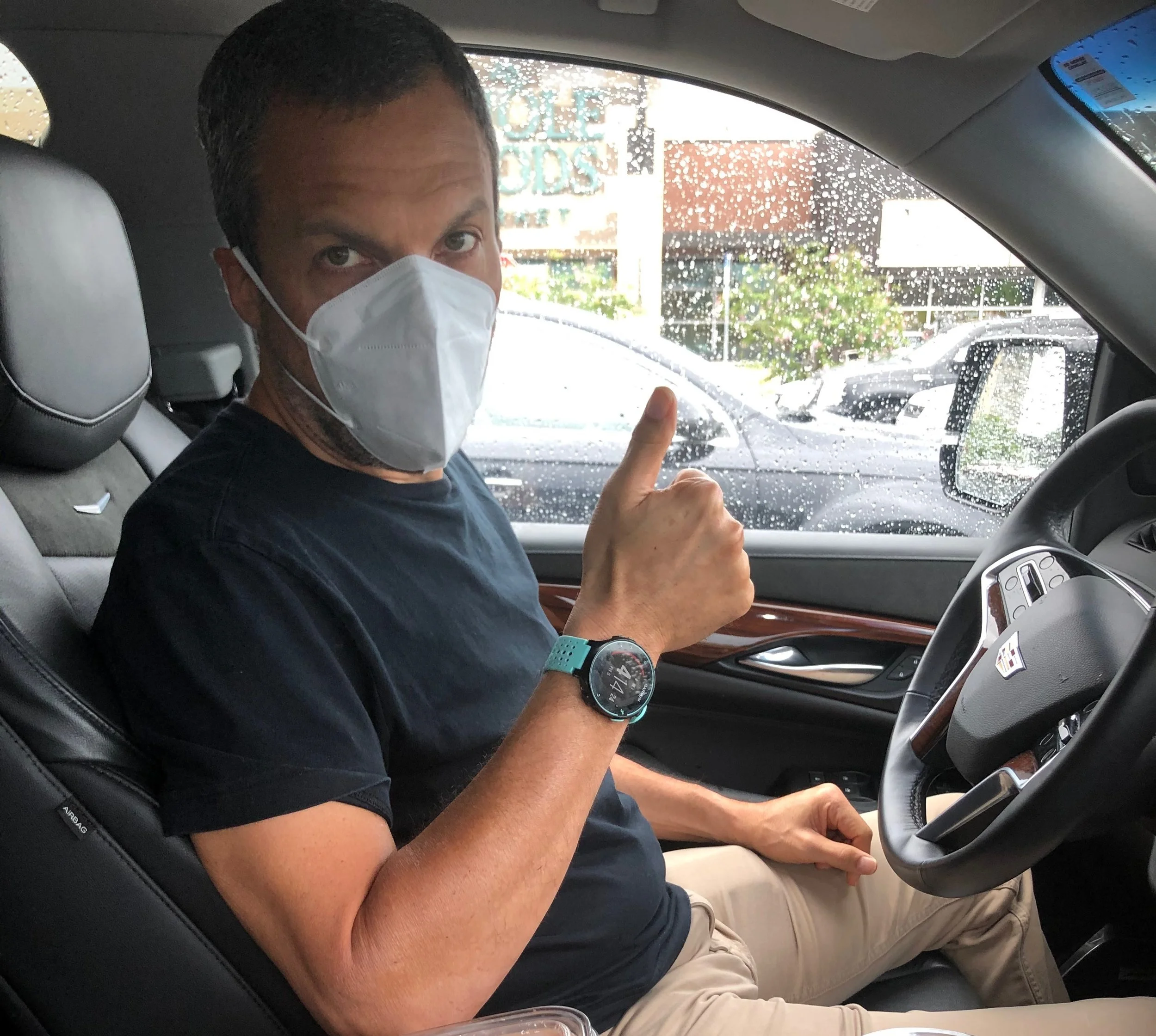 Joel is in the driver's seat of a car with a white mask covering his face. He is giving a thumbs up sign.