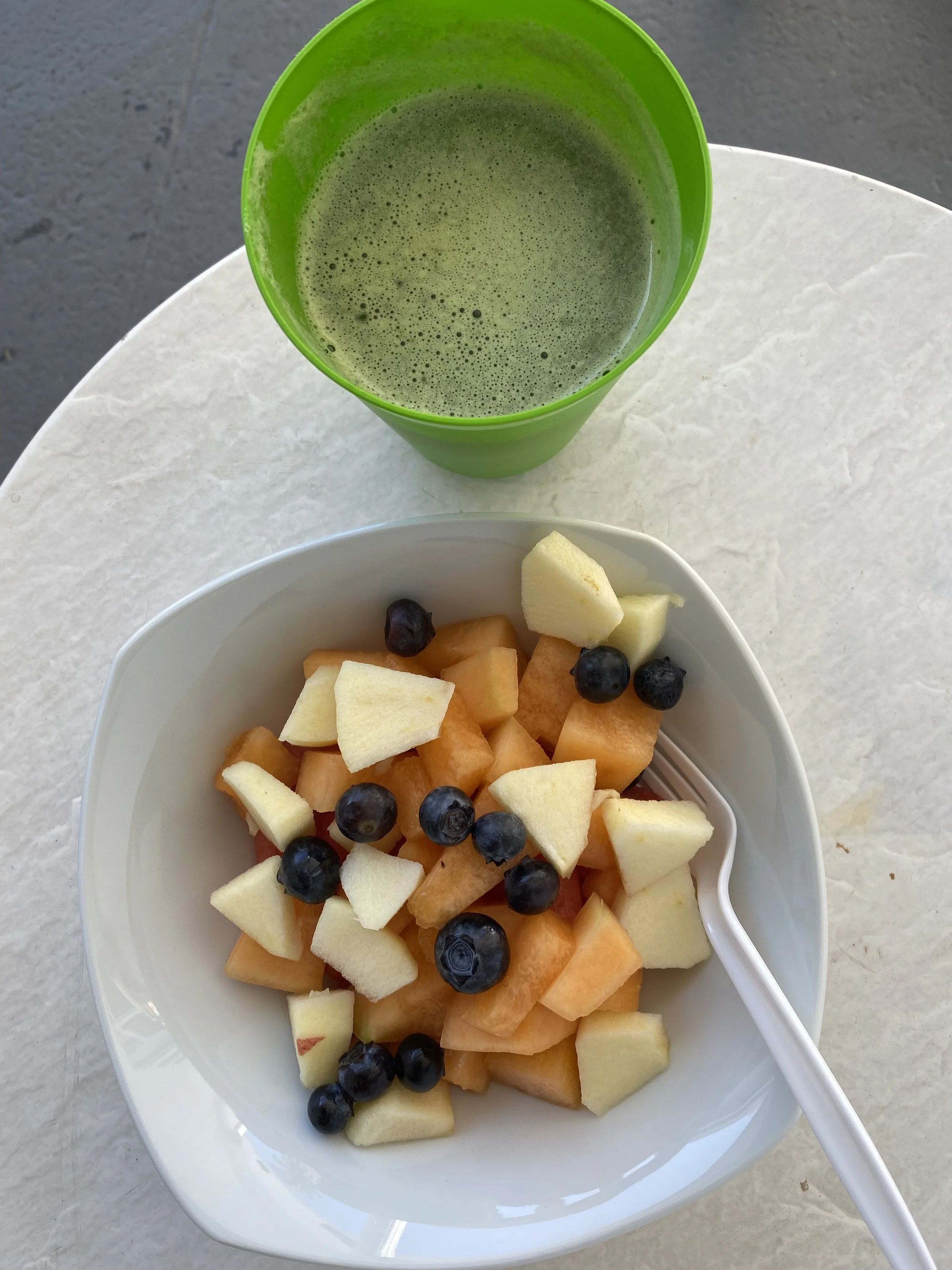 A bowl of fruit (apples, cantaloupe, and blueberries) and a green cup filled with green juice.