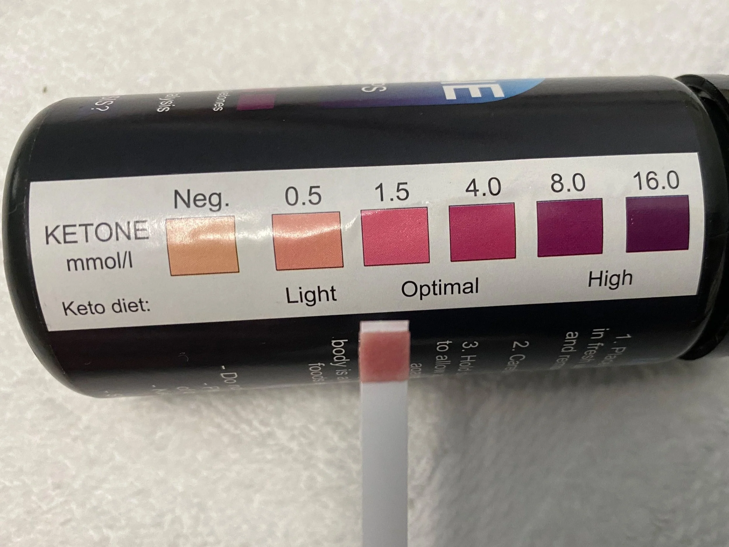 A photo of the measuring scale on the side of a bottle of ketone measuring sticks. There is one stick with a pink tip being held up to the scale measuring an optimal level of ketosis.