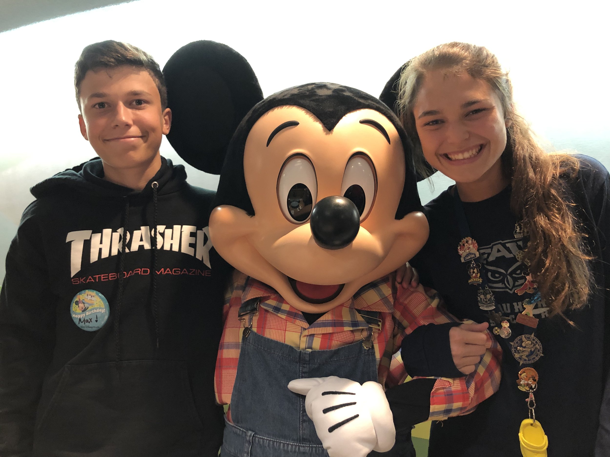 Hannah and Max are pictured with Mickey Mouse. The kids are both wearing black sweatshirts, and Mickey is wearing a plaid shirt with blue overalls.