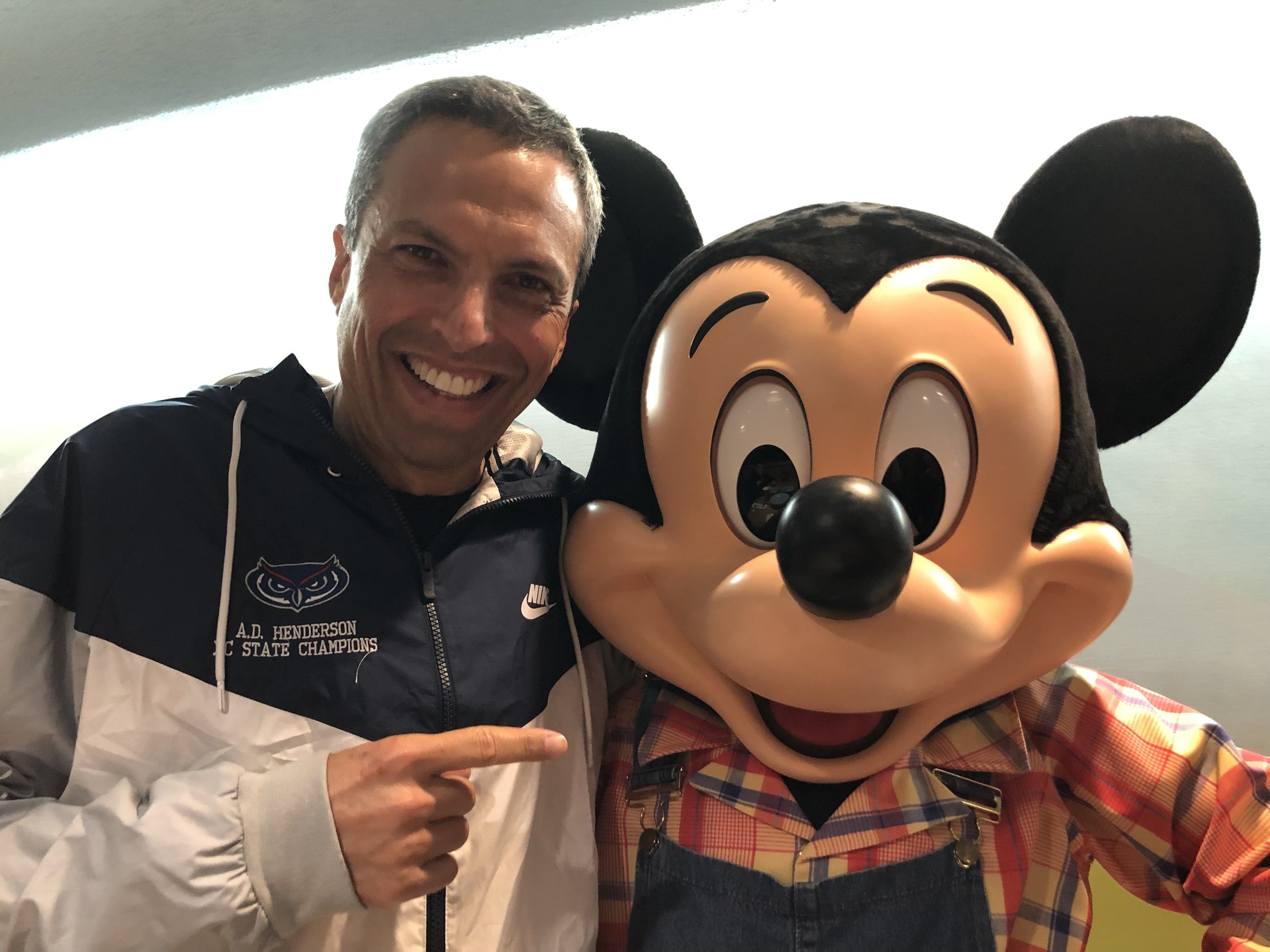 Joel is wearing a blue and white jacket and pointing at Mickey Mouse. Mickey is wearing a plaid shirt with blue overalls.