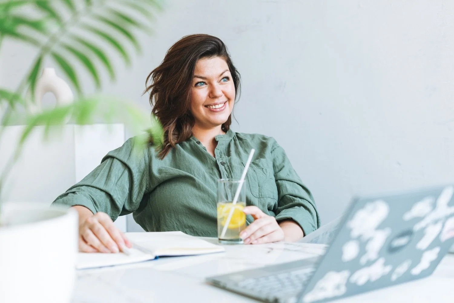 Brunette female wearing a bottle green shirt sits at a desk smiling and holding a glass of water with lemon.